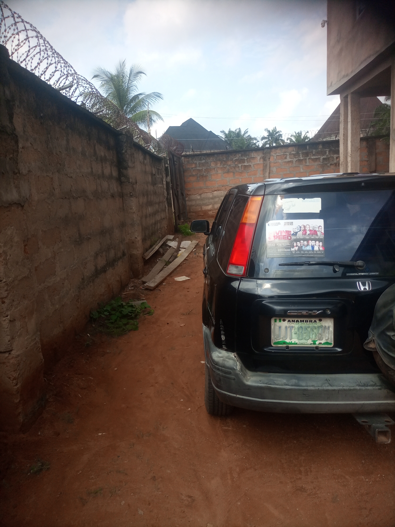 The image captures an outdoor scene in Nkpor, Nigeria, likely during the daytime with fair weather. The foreground is dominated by the rear of a black Honda CR-V, with a license plate reading "JJT-856M" and "ANAMBRA LIGHT OF THE NATION" below it. A sticker featuring a group of people and the text "DIVINE SPEED" is affixed to the rear window. To the left of the car, a rough, weathered brick wall runs along the scene, topped with barbed wire, suggesting a secure or private area.
The ground is a reddish-brown dirt, with some sparse green vegetation growing near the wall and in the path. A few wooden planks are propped against the wall, indicating potential ongoing construction or storage. In the background, more brick walls and structures are visible, including palm trees, hinting at a tropical or subtropical environment. The overall impression is of a functional, somewhat utilitarian outdoor space within a built-up area. There are no explicit clues about the time of day beyond the general brightness, and the weather appears clear. The scene does not convey any specific emotions, presenting a factual depiction of the environment and the vehicle.