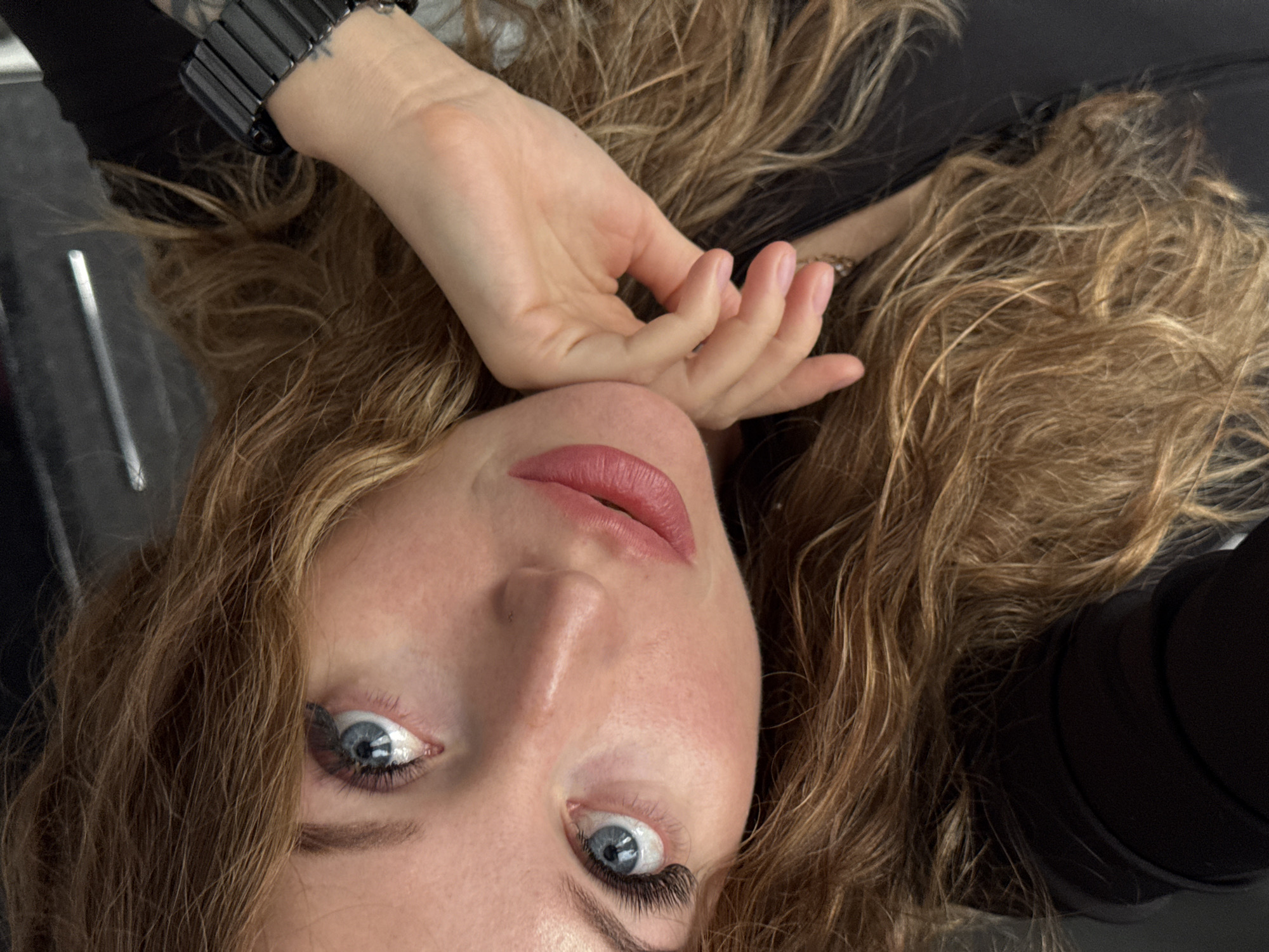 A close-up, tilted view shows a fair-skinned woman with light brown, curly hair spread around her head. She has blue eyes and wears light pink lipstick. Her right hand is gently touching her chin. She wears a black, segmented watch on her left wrist and is dressed in black clothing. The background includes a dark grey surface and part of a silver object. The location is unknown.