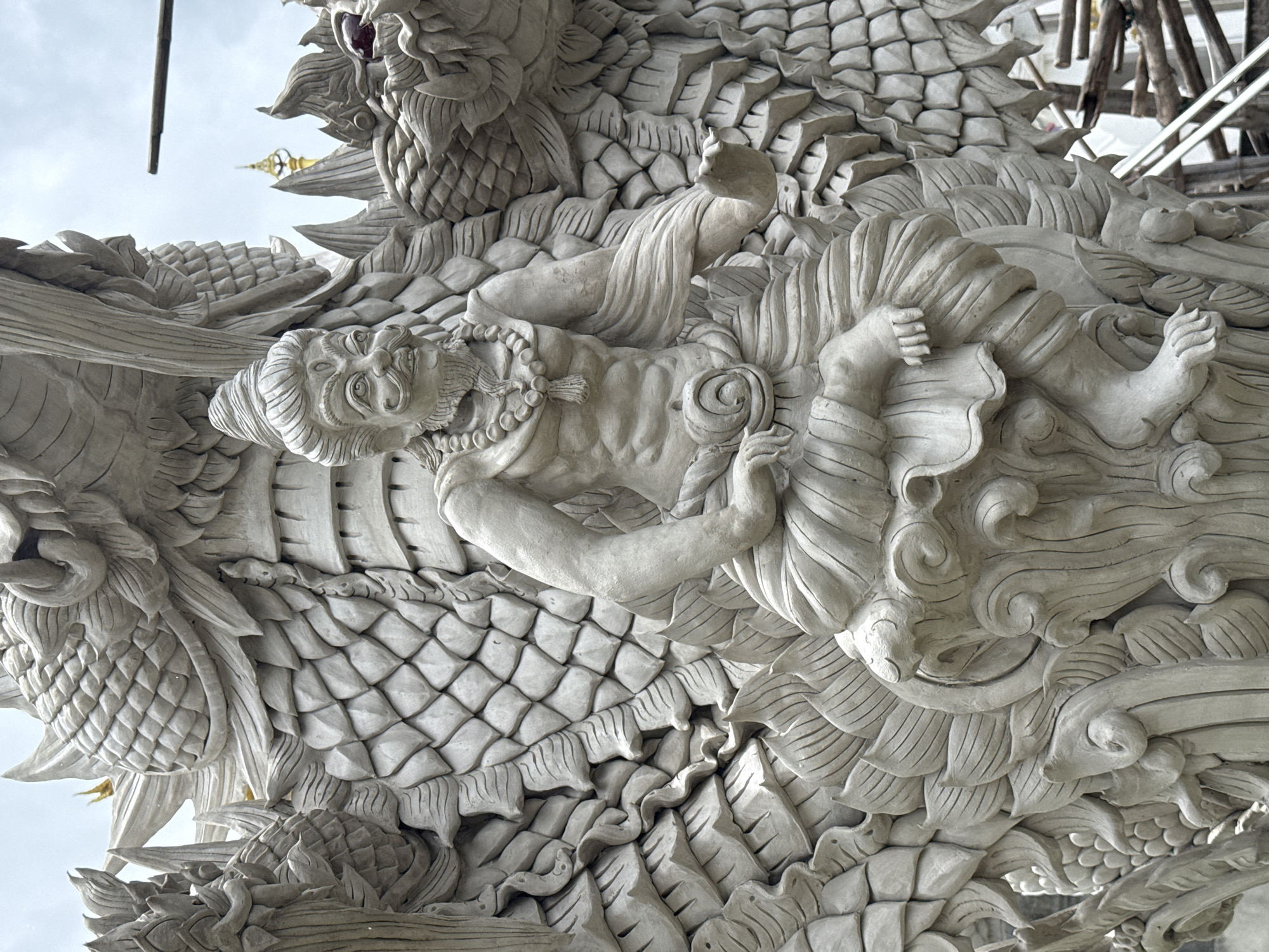 The image shows a close-up view of a large, intricately carved concrete sculpture of a multi-headed Naga serpent, a mythical creature often depicted in Buddhist and Hindu art. A human-like figure, possibly a deity or a sage, is seated in the center of the frame, emerging from the serpent's body. The figure is depicted with a serene expression, adorned with a necklace and a turban. The serpent's scales and undulating form are rendered in great detail, creating a sense of immense power and grandeur. The sculpture is unfinished, with scaffolding visible in the background, suggesting ongoing construction or restoration work. The sky is overcast, and the overall lighting is soft. The location is identified as Ban Sida Ngam, Thailand, which is known for its Buddhist temples and sculptures.