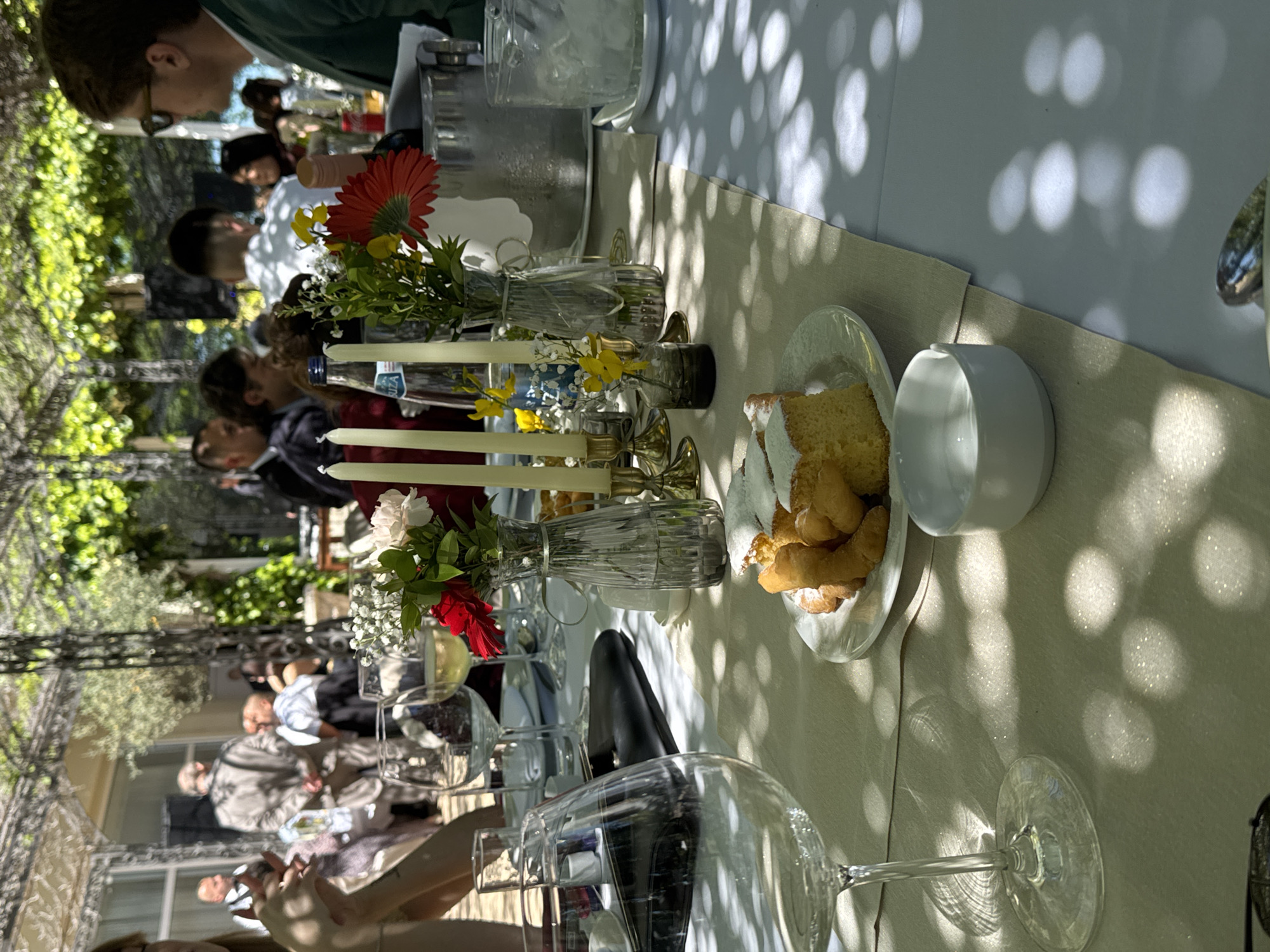 The scene is an outdoor dining arrangement in Petrovac centar, Montenegro. People are seated at a long table covered with a white tablecloth and a beige runner. The table is decorated with floral arrangements in glass vases, candles, and plates of cake and pastries. The light is dappled with shadows from foliage above. Wine glasses are prominent in the foreground. The scene appears to be a meal or gathering in an outdoor restaurant or event space, with people eating and socializing. The surroundings are leafy, with architectural elements of a building visible in the background.