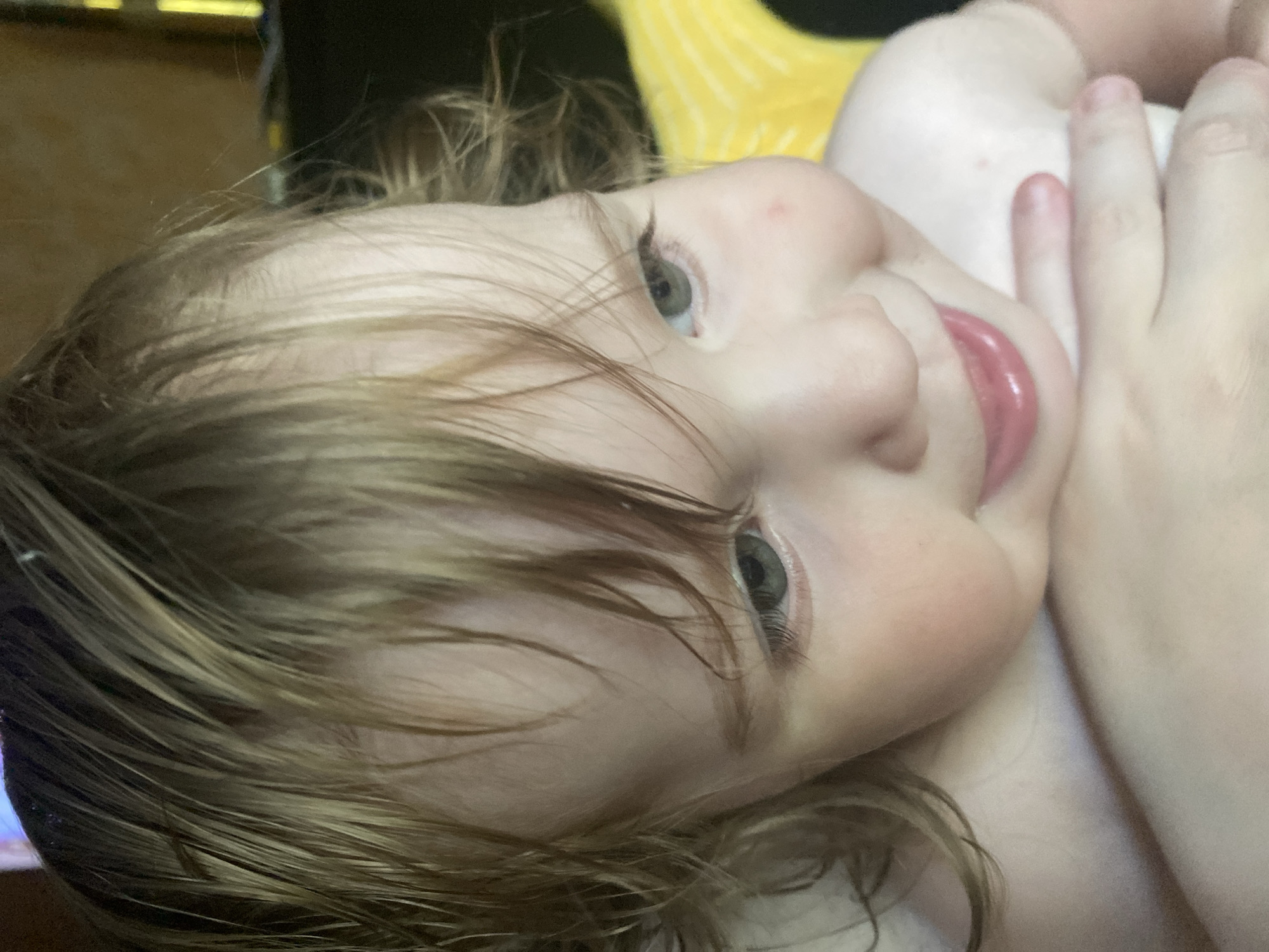 A close-up, tilted, eye-level shot shows a baby's face. The baby has light brown hair, pale skin, and light green eyes. The baby's mouth is slightly open and appears to be smiling. The baby's right hand is visible near its face, with fingers bent. The background is out of focus and appears to be a room with some yellow fabric and dark shapes. The lighting is soft and natural, suggesting daytime. The overall mood is tender and intimate, capturing a moment of innocent joy.