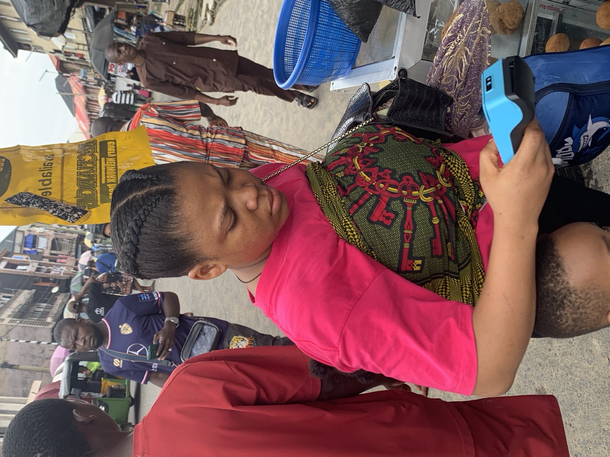 The image depicts a bustling street scene in Aba, Nigeria, likely a market area given the presence of vendors and numerous people. In the foreground, a woman with braided hair is holding a blue and black point-of-sale device, suggesting a commercial transaction is underway. She is wearing a vibrant pink t-shirt and a patterned fabric wrapped around her waist. Behind her, a man in a purple t-shirt with a logo is looking to his left, holding a mobile phone. Other individuals are visible in the background, some holding umbrellas, indicating overcast weather or protection from the sun. The environment appears to be a crowded urban setting with buildings lining the street and various goods displayed on tables. The overall impression is one of lively commerce and daily life in a Nigerian city.