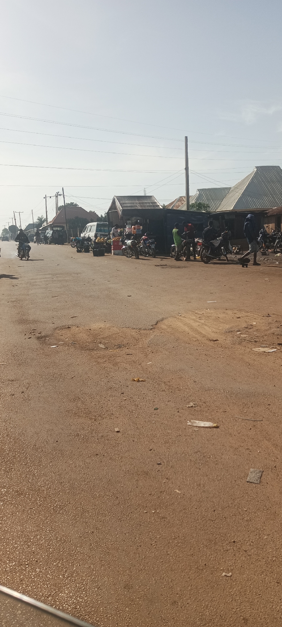 This image depicts a bustling street scene under bright daylight. The foreground features a wide road, a mix of paved asphalt and reddish-brown dirt, with several potholes and loose debris, including a plastic bottle and wrappers.
In the midground and background, numerous people are visible, though their features are indistinct. On the left, a person rides a motorbike away from the viewer. On the right side of the street, a market or commercial area is active. Several individuals are gathered near makeshift stalls and shops, constructed with simple frames and corrugated metal roofs. Merchandise, notably piles of fruits such as bananas and oranges, is displayed outside these stalls.
A significant number of motorbikes are parked or standing near the stalls, suggesting they are either customer vehicles or used for local transport. A white van and another dark-colored vehicle are also parked further down the road. Overhead, power lines crisscross the sky, supported by wooden utility poles, indicating electrical infrastructure. The sky is a bright, clear light blue, suggesting dry weather. The overall atmosphere points to a busy, everyday environment in a potentially rural or semi-urban setting. No legible text is visible in the image. The bright, even lighting indicates it is likely mid-morning or early afternoon.