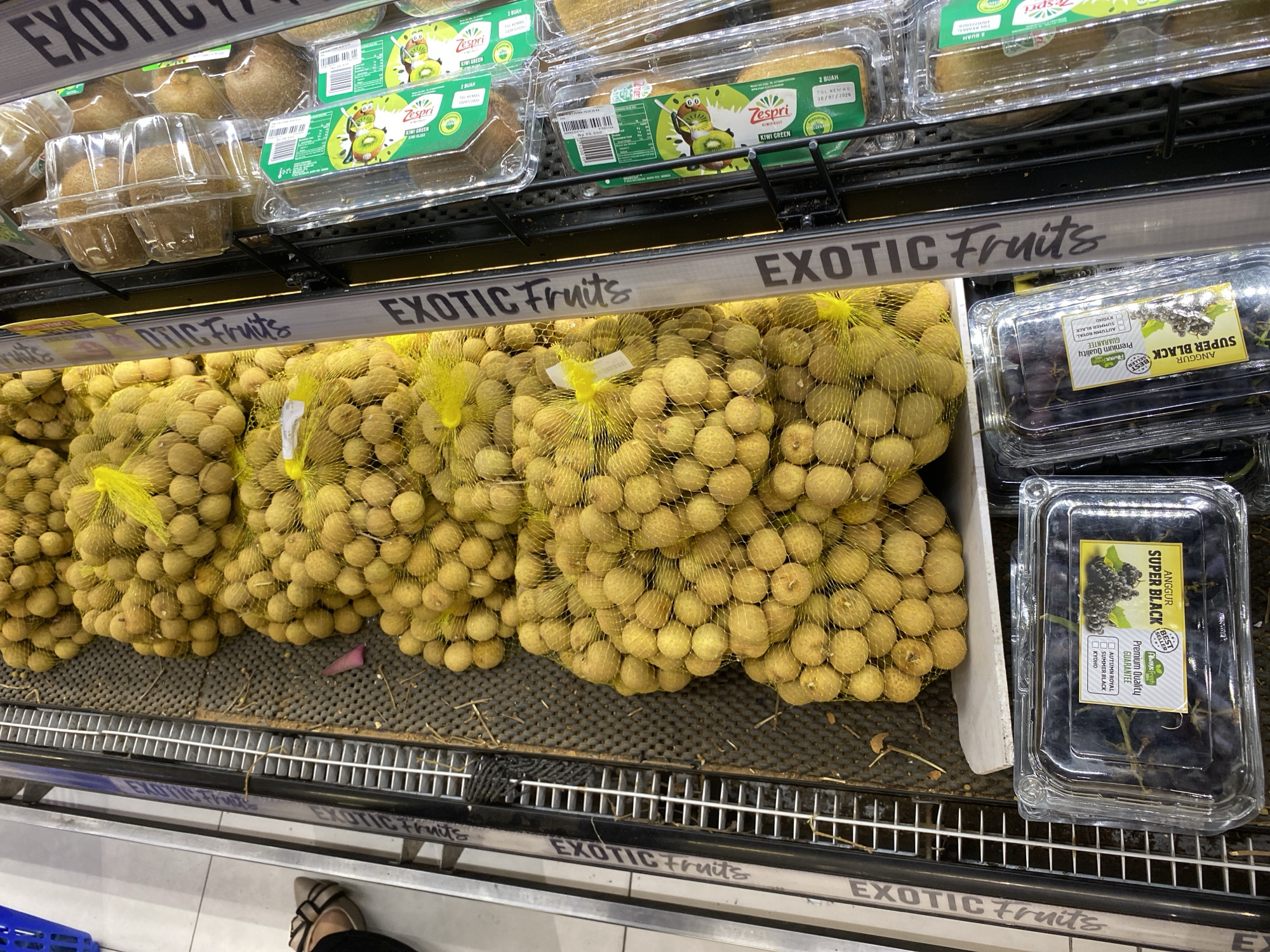 The image shows a display of fruits in a supermarket refrigerator section in Banda Aceh, Indonesia. In the foreground, there are several clear plastic containers of kiwi fruit, some with green packaging. Above the kiwis, a shelf displays bags of longans, which are small, round, yellow-skinned fruits, contained within yellow netting. To the left of the longans, there is a clear plastic container of dark purple grapes, labeled "ANGGUR SUPER BLACK." A sign above the fruit display reads "EXOTIC FRUITS." A person's legs and feet are partially visible in the upper right corner of the image, wearing beige shoes. The environment appears to be a well-lit retail space with metal shelving and a tiled floor.
