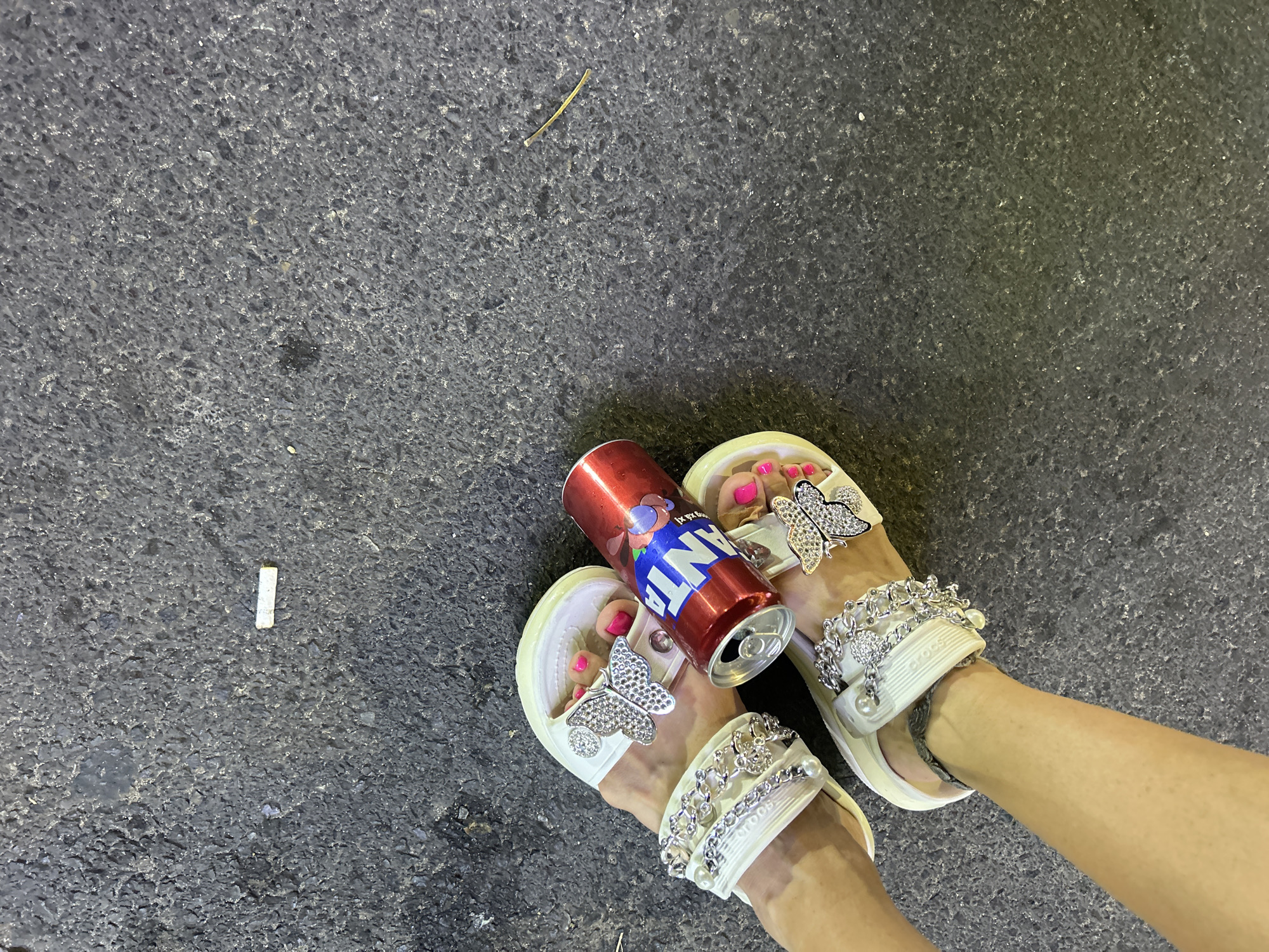 The image shows a pair of feet wearing white Crocs with rhinestone butterfly decorations. The toenails are painted bright pink. An open can of red Fanta soda sits between the feet. The feet are on a dark gray paved surface with a cigarette butt lying to the left of the feet. The specific location in Nha Trang, Vietnam cannot be determined from this image alone.