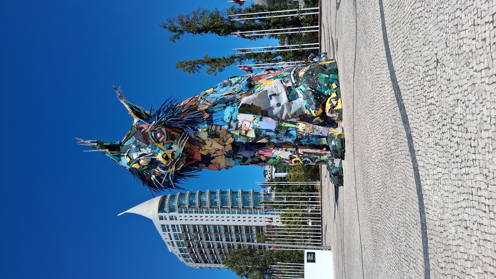 In Lisbon, Portugal, a large, colorful sculpture of a lynx stands in a public plaza under a clear blue sky. The sculpture, made from recycled materials, is the focal point of the scene. Behind the sculpture, a modern, white, high-rise building with a distinctive curved top rises above other structures. In the middle ground, a series of white poles topped with various national flags are arranged in a line, suggesting an international or diplomatic setting. Trees dot the landscape, and a few people are visible in the distance, some seated and others walking. The ground of the plaza is paved with light-colored cobblestones.