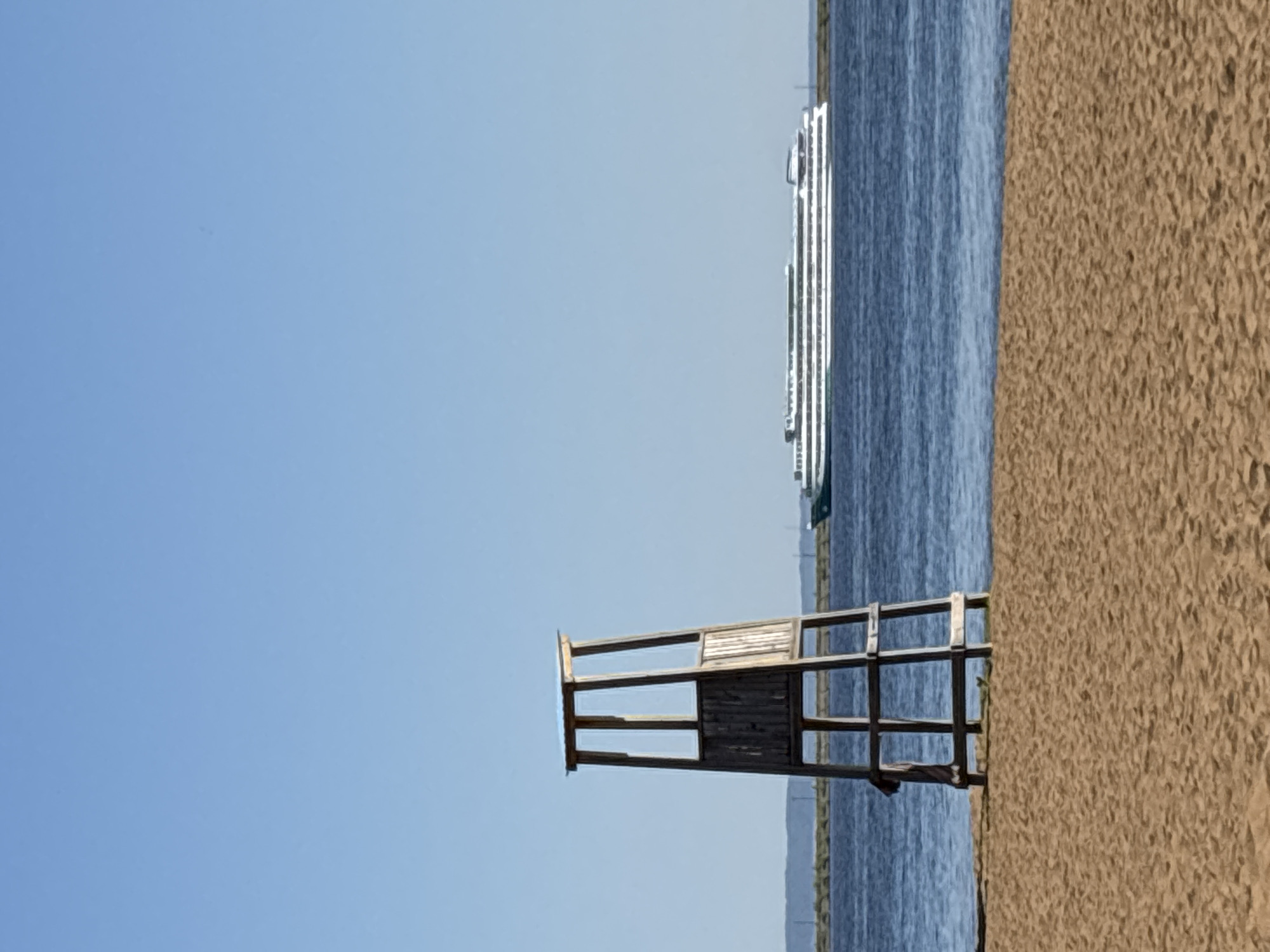 The image depicts a beach scene. In the foreground, there is a wooden lifeguard chair positioned on the sandy beach. Beyond the beach, there is a body of water, possibly a river or lake, and a cruise ship is visible in the distance. The sky is clear and blue. The location is Chaikovsky, Russia.