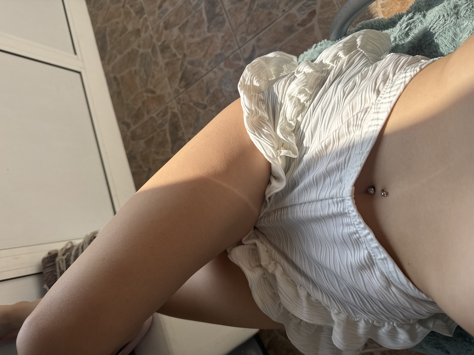 An overhead, close-up shot captures a person's lower torso and upper legs while they are resting or lounging. The subject wears a white, crinkled or pleated garment, resembling shorts or a ruffled skirt, which partially covers their midsection and upper thighs. A silver belly button piercing, a barbell style with two balls, is visible above the waistband of the clothing.
Strong sunlight illuminates the scene, casting a clear tan line across the upper left thigh and creating bright highlights on the skin and fabric. The skin appears sun-kissed.
The background features a tiled floor with earthy brown and grey tones, typical of indoor or outdoor paving. A white door frame or sliding door is visible on the left side of the frame. A soft, teal-colored fluffy fabric, possibly a blanket or cushion, can be seen beneath the person's upper body, just out of frame. A small, dark, woven object is partially visible near the bottom left by the door frame.
The image conveys a relaxed atmosphere on a bright, sunny day, likely in an indoor setting or on a balcony/patio. There is no visible text.