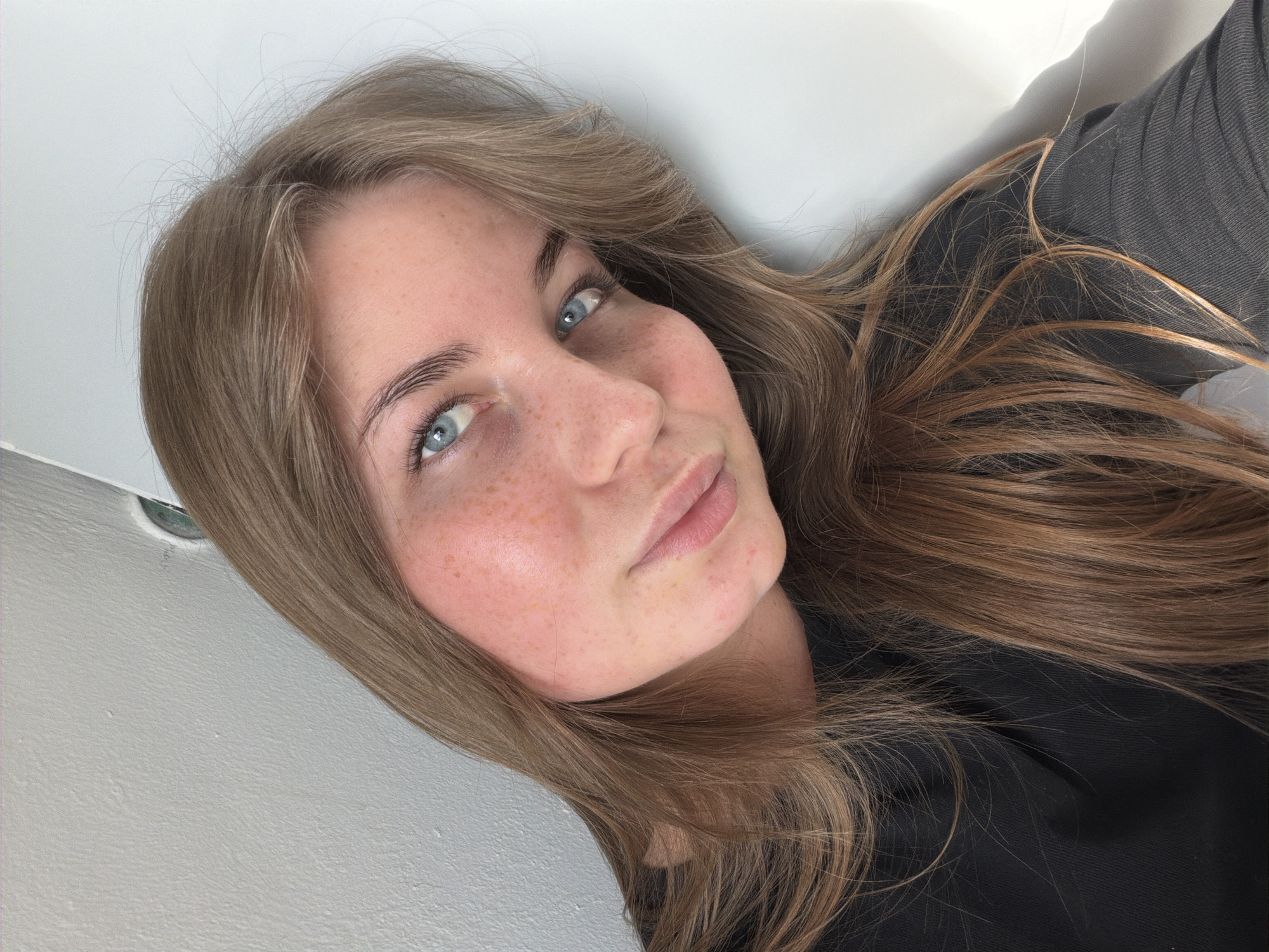 A young woman with blonde hair and blue eyes, wearing a black shirt, leans her head against a white wall. She has freckles on her face and her lips are slightly parted. The lighting suggests an indoor setting, likely a room with natural light coming from the right. The image is a close-up portrait, focusing on her face and upper body. There are no visible landmarks or features that would indicate a specific location in Chaikovsky, Russia.