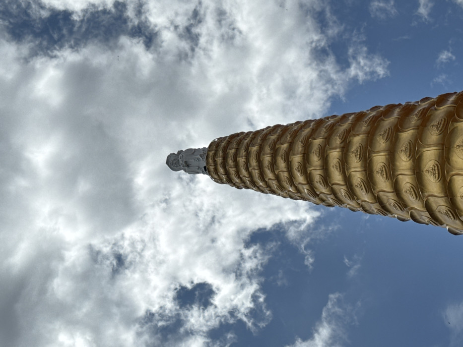 The image depicts a tall, multi-tiered religious structure, likely a stupa or pagoda, located in Đăk Mar, Vietnam. The structure is golden in color, conical, and tapers significantly upward. Each tier features multiple relief carvings of seated figures, presumably Buddhas, in a meditative posture. At the apex of this golden structure stands a distinct, lighter-colored statue, possibly white or light grey. This statue portrays a robed figure, identifiable as a deity or Bodhisattva, with an elaborate headpiece or hairstyle. The scene is set against a partly cloudy sky, showing patches of clear blue sky interspersed with white and grey cumulus clouds, indicating daylight conditions. No other objects or individuals are present.