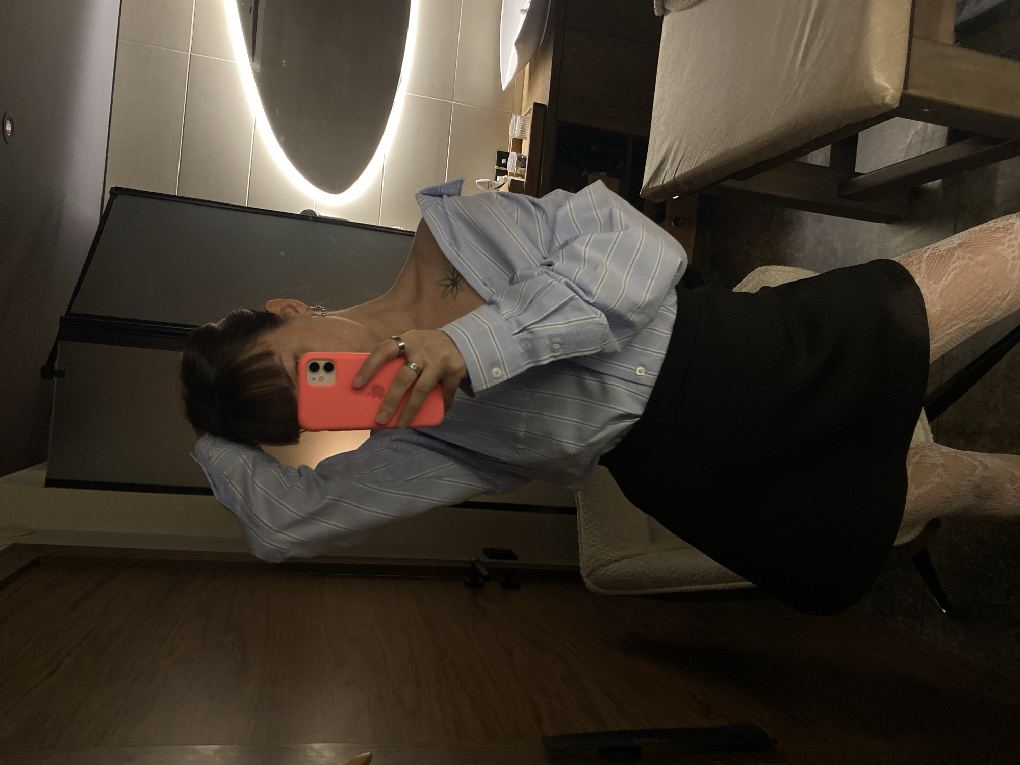 A person is taking a selfie in a room. The individual is wearing a blue and white striped shirt, a black skirt, and white patterned tights. They are holding a pink phone. The background includes a lit, rounded mirror, tiled walls, a dark screen, wooden flooring, and furniture. The location is unknown.