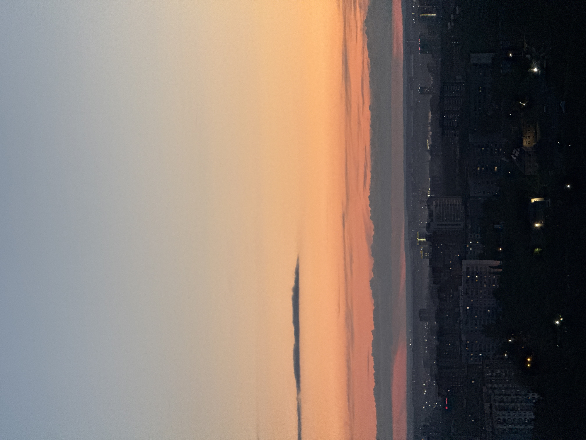 The image displays a panoramic twilight scene over the city of Perm, Russia, likely captured during sunset from an elevated vantage point. The horizon divides the frame, with the sky occupying the upper portion and the urban landscape below.
The sky exhibits a beautiful gradient of colors, transitioning from a soft, muted blue-grey on the left to vibrant shades of peach, orange, and soft pink towards the right. Several long, thin, dark cloud formations stretch horizontally across the warmer parts of the sky, with one prominent dark cloud appearing as a distinct streak in the central area.
Below the colorful horizon, the sprawling city of Perm is enveloped in the dim light of dusk. Buildings are largely silhouetted against the fading light, but numerous windows are illuminated, and streetlights create a scattered pattern of bright points across the dark urban canvas. The artificial lights suggest the city is transitioning into night.
No people or specific activities are visible, contributing to a serene and contemplative atmosphere. The contrast between the radiant, colorful sky and the dark, light-dotted cityscape is a notable detail. The image has a slightly grainy texture, characteristic of low-light photography. No visible text is present in the image.