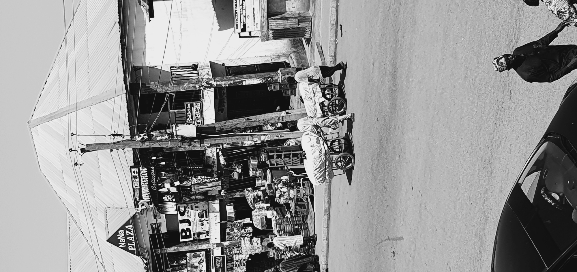 A black and white photograph captures a street scene in Bukuru, Nigeria, with a focus on a bustling marketplace and people engaged in daily activities. In the background, a multi-story building with shops displays signage, including "NaNana PLAZA" and "Phenomenon Collection." Goods are displayed outside the shops, with clothing items hanging from racks and various items stacked on shelves and tables.
In the foreground, two men are pushing carts laden with covered goods along a paved road. The carts are metal-framed with spoked wheels. One man, wearing light-colored clothing, is positioned behind the cart on the left, while the other man, in darker attire, pushes the cart on the right. To the left of the scene, a person in a white striped robe is walking away from the camera, and further left, a portion of a black car is visible. The overall environment appears to be a sunny day, indicated by the shadows cast on the ground. The pavement shows some wear and tear.