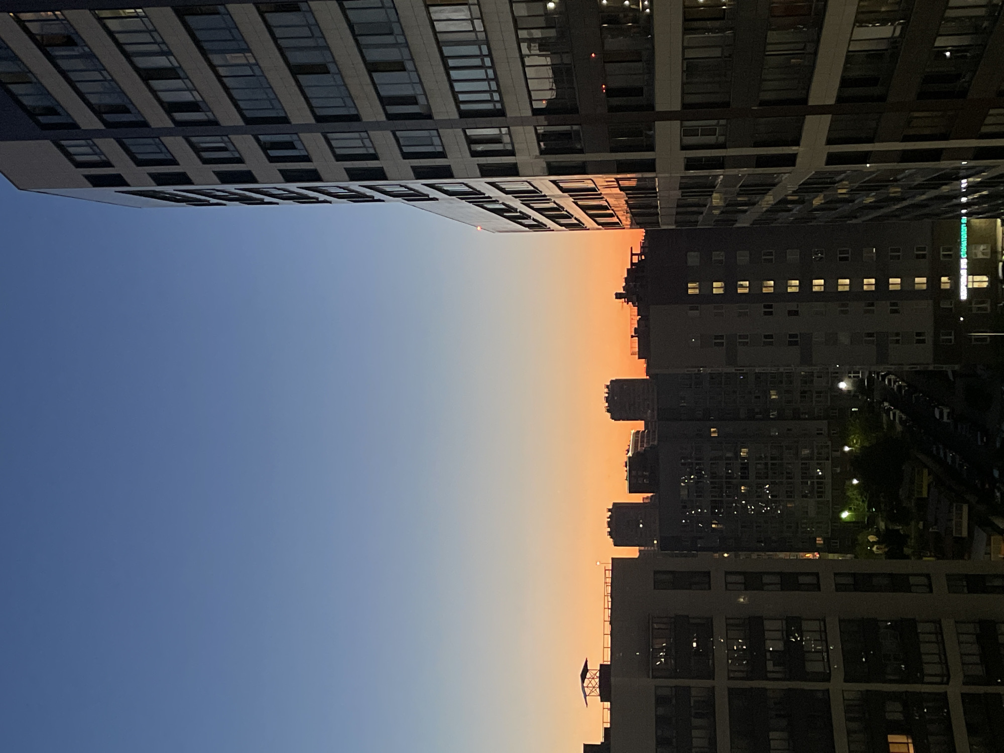 This is a vertical, wide shot of an urban landscape during dusk. The scene is dominated by large, modern apartment buildings, some with extensive window facades that reflect the sky, while others show illuminated windows indicating occupancy. The buildings are arranged on both the left and right sides of the frame, creating a canyon-like effect.
The sky transitions from a deep blue at the top of the frame to a vibrant orange-pink near the horizon, characteristic of sunset. Silhouetted against this colorful sky are more apartment buildings in the distance. The overall impression is one of a densely populated urban environment at the end of the day.
There are no people visible in the frame. The objects are primarily the architectural structures of the apartment buildings, with their geometric lines and numerous windows. The setting is clearly a modern city or a large residential complex.
The time of day is evident from the gradient of the sky, indicating sunset. The weather appears clear, with no clouds. There are no explicit location cues, but the mention of Sinyavino, Russia, provides geographical context. The image evokes a sense of calm and the transition from day to night in an urban setting.
No visible text is present in the image.