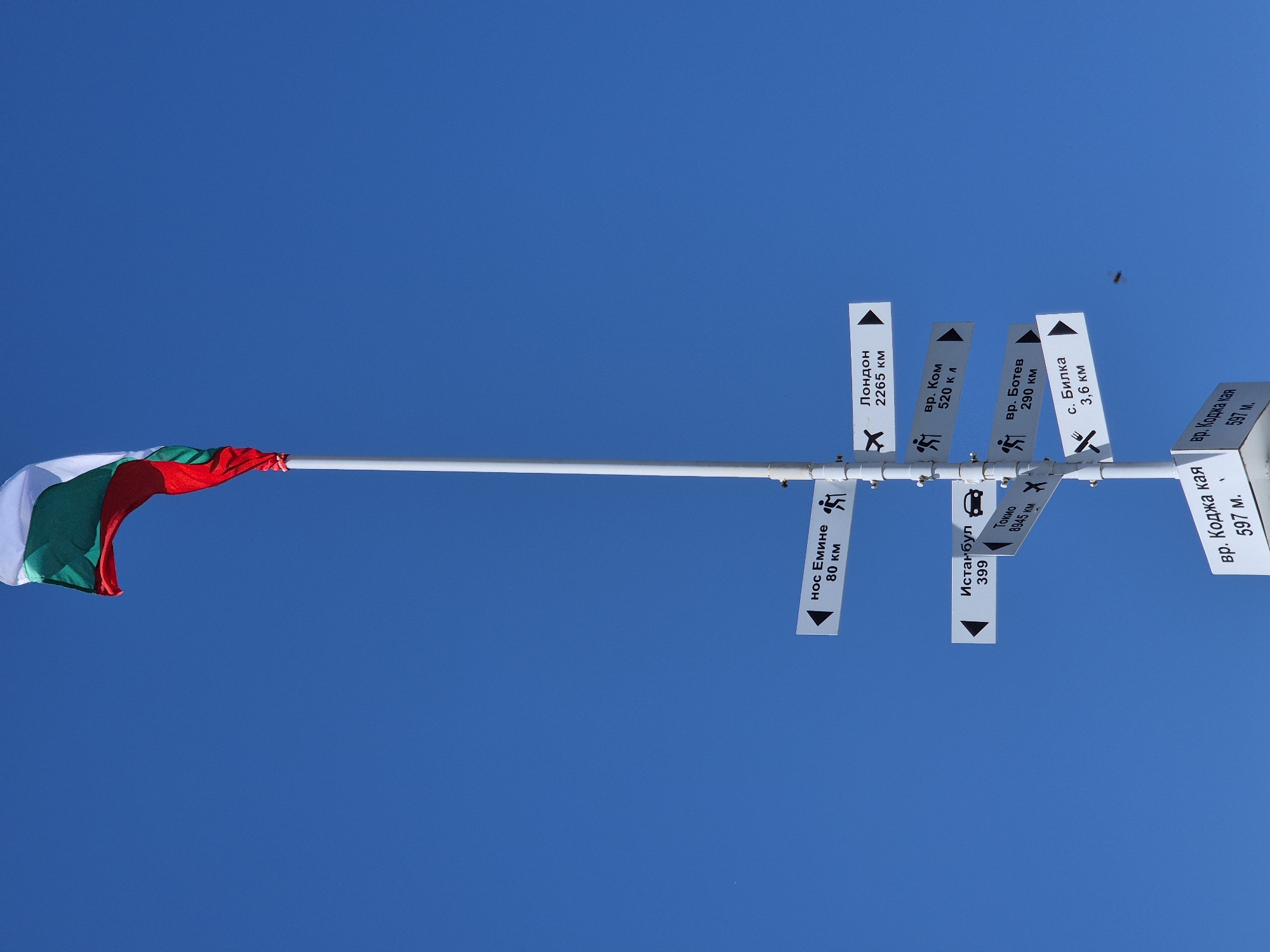 The image shows a Bulgarian flag attached to a white pole, which also supports several direction signs pointing in different directions. The background is a clear, bright blue sky. The signs indicate directions to various places and their distances: London (2265 km), vr. Kom (520 km), vr. Botev (290 km), the village of Bilka (3.6 km), vr. Kodja Kaya (597 m), Nos Emine (80 km), Istanbul (399 km), and Tokyo (8945 km). The presence of a direction sign pointing to the village of Bilka suggests that this photograph was taken near or in Bilka, Bulgaria. The signs feature symbols indicating if the destinations are for hikers or drivers.