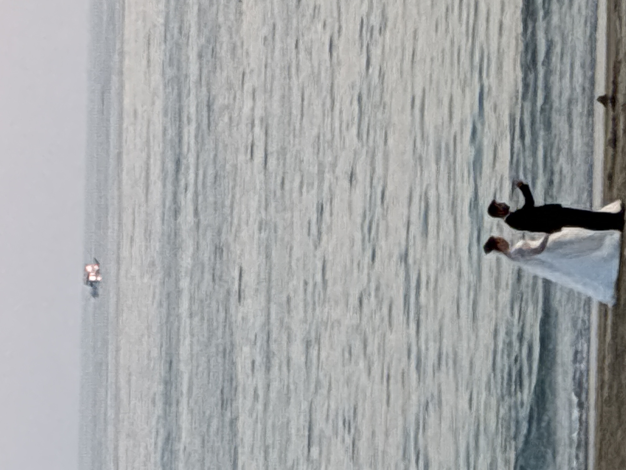 An out-of-focus, medium shot depicts a wedding couple on the shore of the sea, with a distant boat in the background. The man is on the right, wearing a dark suit and white shirt, with his right arm extended as if pointing. The woman is on the left, wearing a white wedding dress with a train that extends onto the sand. The sea is visible behind them, with choppy water and a slightly hazy sky. The beach is sandy and appears to be a natural setting. The boat in the distance appears to be a small vessel with bright lights. The overall scene suggests a wedding photoshoot or a moment captured on a beach, possibly in Alanya, Turkey, given the context of the provided information.