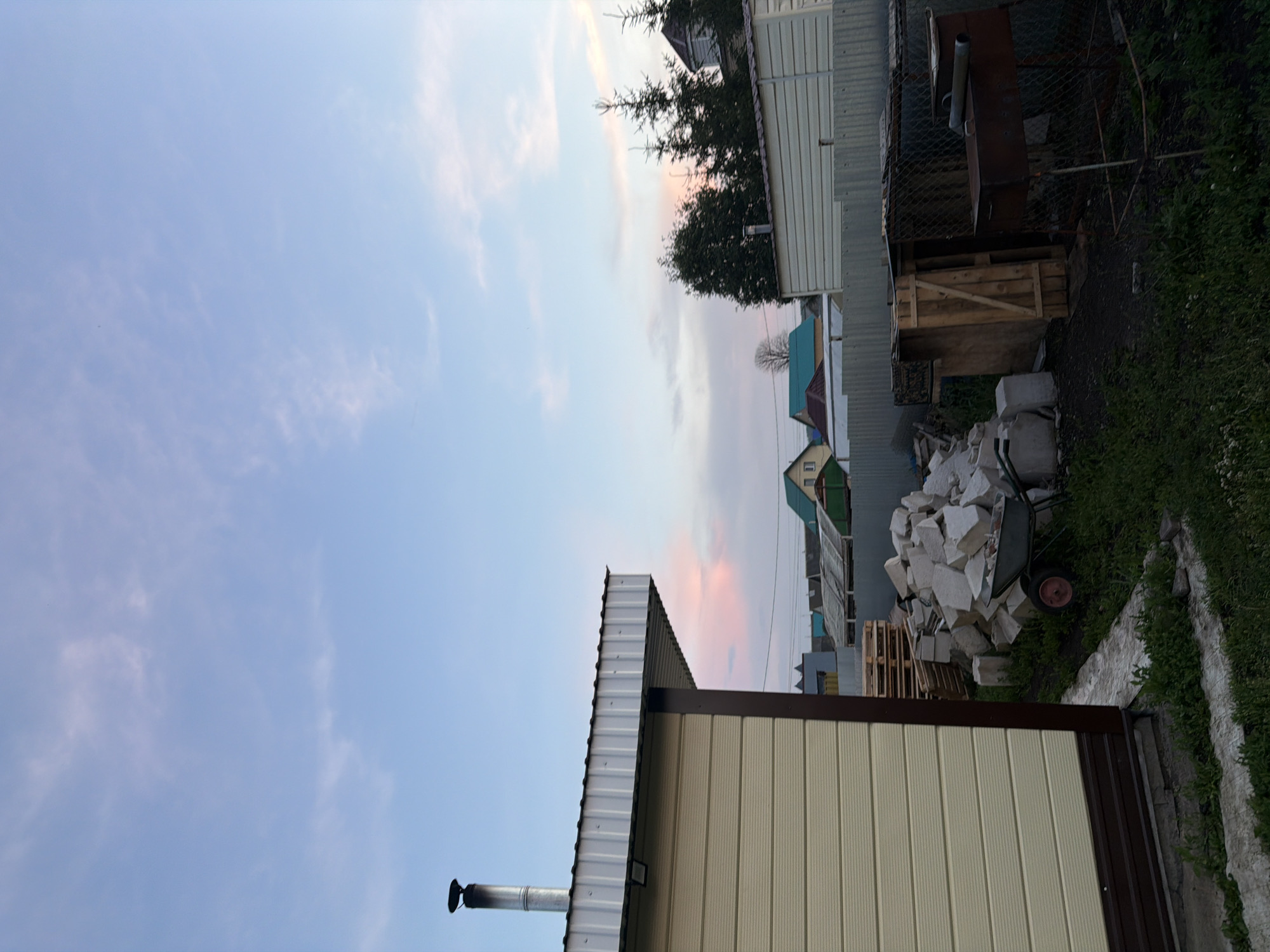 The image depicts a backyard scene in Nizhny Suyuk-Su, Russia, during what appears to be late afternoon or early evening, given the soft lighting and pastel hues in the sky. In the foreground to the left is the side of a light-colored, corrugated building with a dark brown trim along the base and corner. A metal chimney extends vertically from the roof of this building. Adjacent to this structure, a pile of light-colored rubble, possibly construction debris, is present, with a wheelbarrow to the left of the pile. Behind the rubble, a wooden shed with a slanted roof and a door made of vertical planks is visible. To the right of the shed, a rusty barbecue grill with two cylindrical pipes on top is set up on legs and enclosed by a chain-link fence. In the background, a series of houses with various colored roofs, including green and red, are scattered across the landscape. Pine trees are visible behind the houses. The sky is a soft blue with wispy, pink-tinged clouds. The ground is covered with grass and some concrete pathways or slabs. There are no people visible in the image.