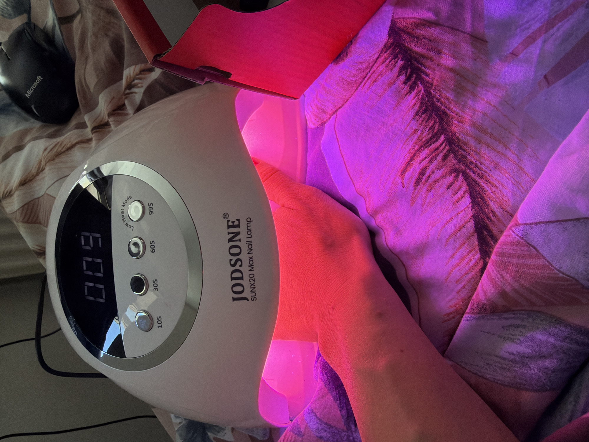 The image shows a hand inside a JODSONE SUNX20 Max Nail Lamp. The lamp is turned on and displaying "009" on its digital timer. The buttons for "Low Heat Mode," "10S," "30S," "60S," and "99S" are visible on the lamp. Next to the lamp is a Microsoft computer mouse. The background includes a patterned surface that looks like a blanket or bedding. There is no verifiable evidence in the image to associate the scene with the city of Bearpark, United Kingdom.