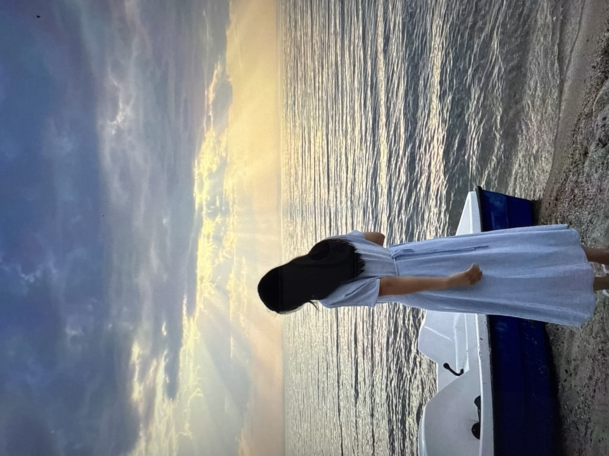 The image shows a young woman with long, dark hair standing on a beach, looking out at the sea. She is wearing a light blue dress. Next to her is a white and blue paddleboat. The sky is cloudy, with rays of sunlight breaking through the clouds. The sea is calm with small waves. The location is identified as Găvănoasa, Moldova.