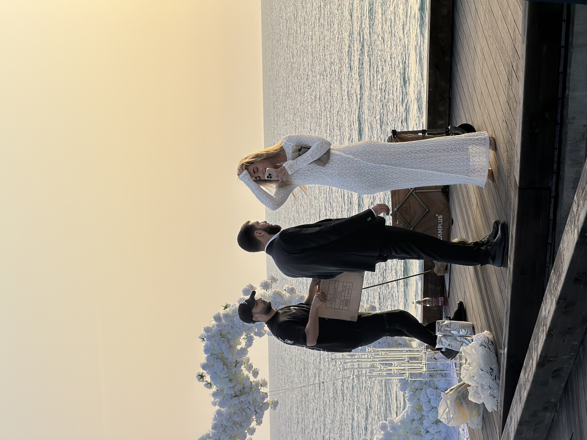 On a wooden pier jutting out into the sea, three people are visible. A woman in a white dress holds a mobile phone to her head. Next to her stands a man in a suit. Another man, wearing a black baseball cap and black clothes, carries a cardboard box. The pier appears to be the site of a wedding setup, as there is a white floral arch present. The location is Bat Yam, Israel, judging from known locations and geographical features.