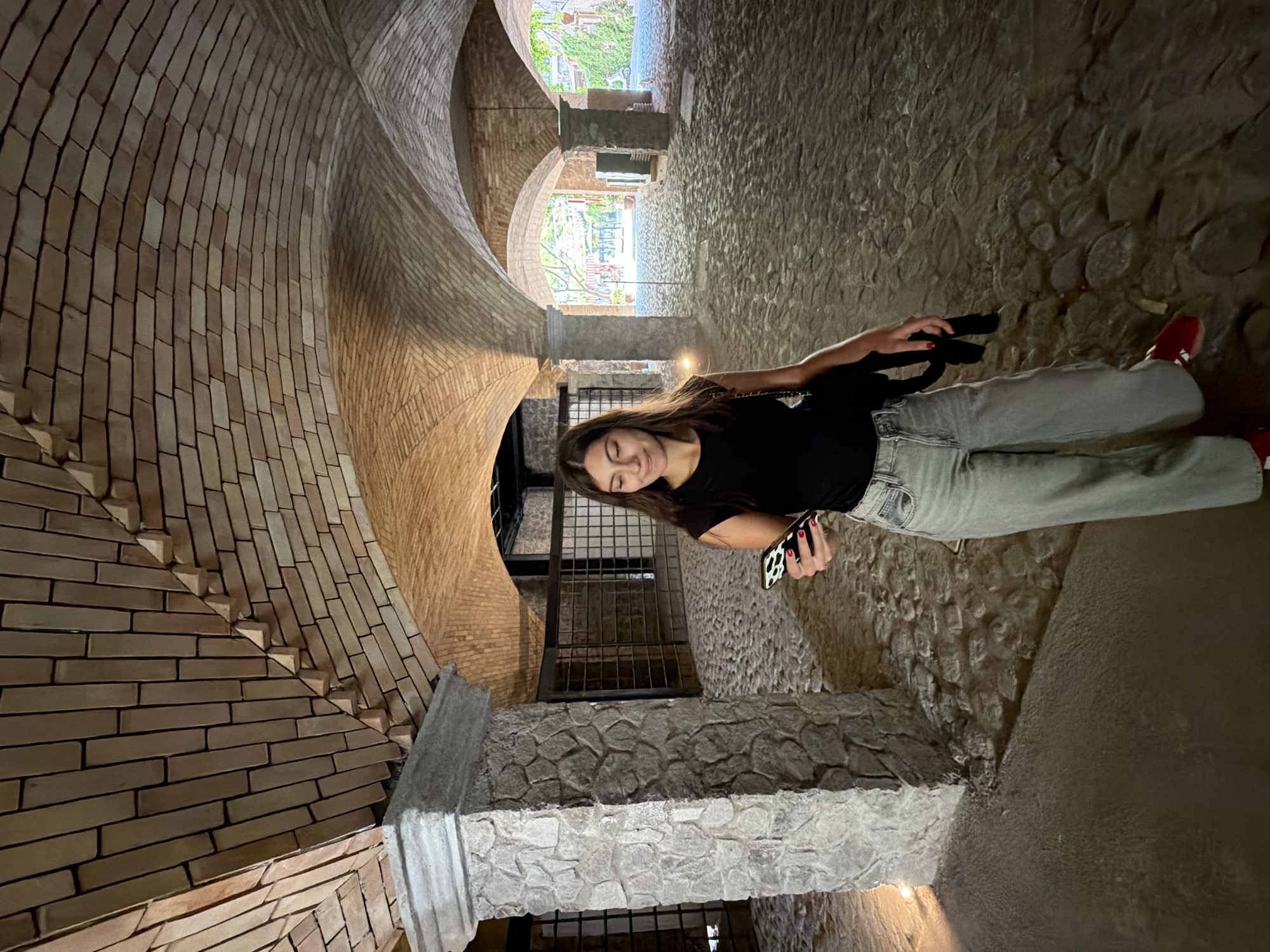 A young woman stands in an arched tunnel in Domingo Arenas, Mexico, looking at her phone. She is dressed in a black shirt, light gray jeans, and red shoes. The tunnel is constructed of red brick and stone and has a series of arches leading to an opening with visible foliage and buildings in the distance. The tunnel's walls curve dramatically, and the ground is made of cobblestones. There is ironwork visible at various points in the background. The lighting within the tunnel is dim and shadowed.