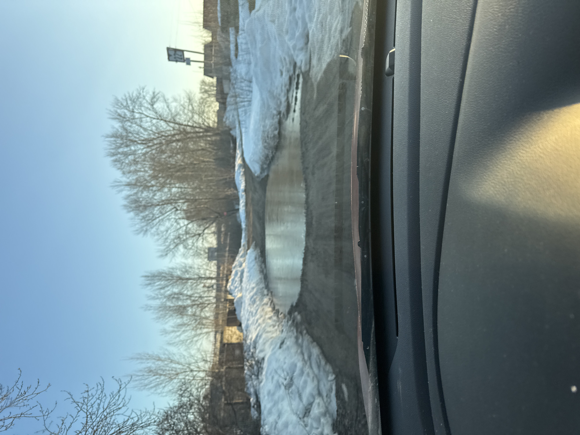 A street in Chaikovsky, Russia, is viewed through a car windshield. A puddle of water fills the road, with patches of melting snow along the sides. Trees with bare branches line the street. A signpost is visible in the distance. The sky is blue. The interior of the car is partially visible.