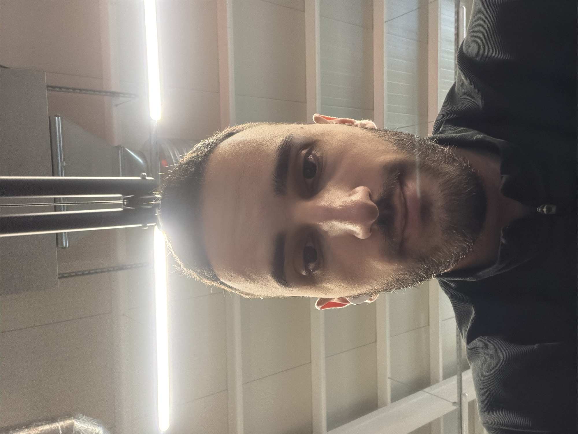 A close-up, horizontally rotated selfie of a light-skinned man with dark hair and a beard, likely indoors under fluorescent lighting. The background appears to be a warehouse or industrial-style ceiling with visible support beams and strip lighting. No specific identifying markers are present to definitively locate the scene within Plovdiv, Bulgaria.