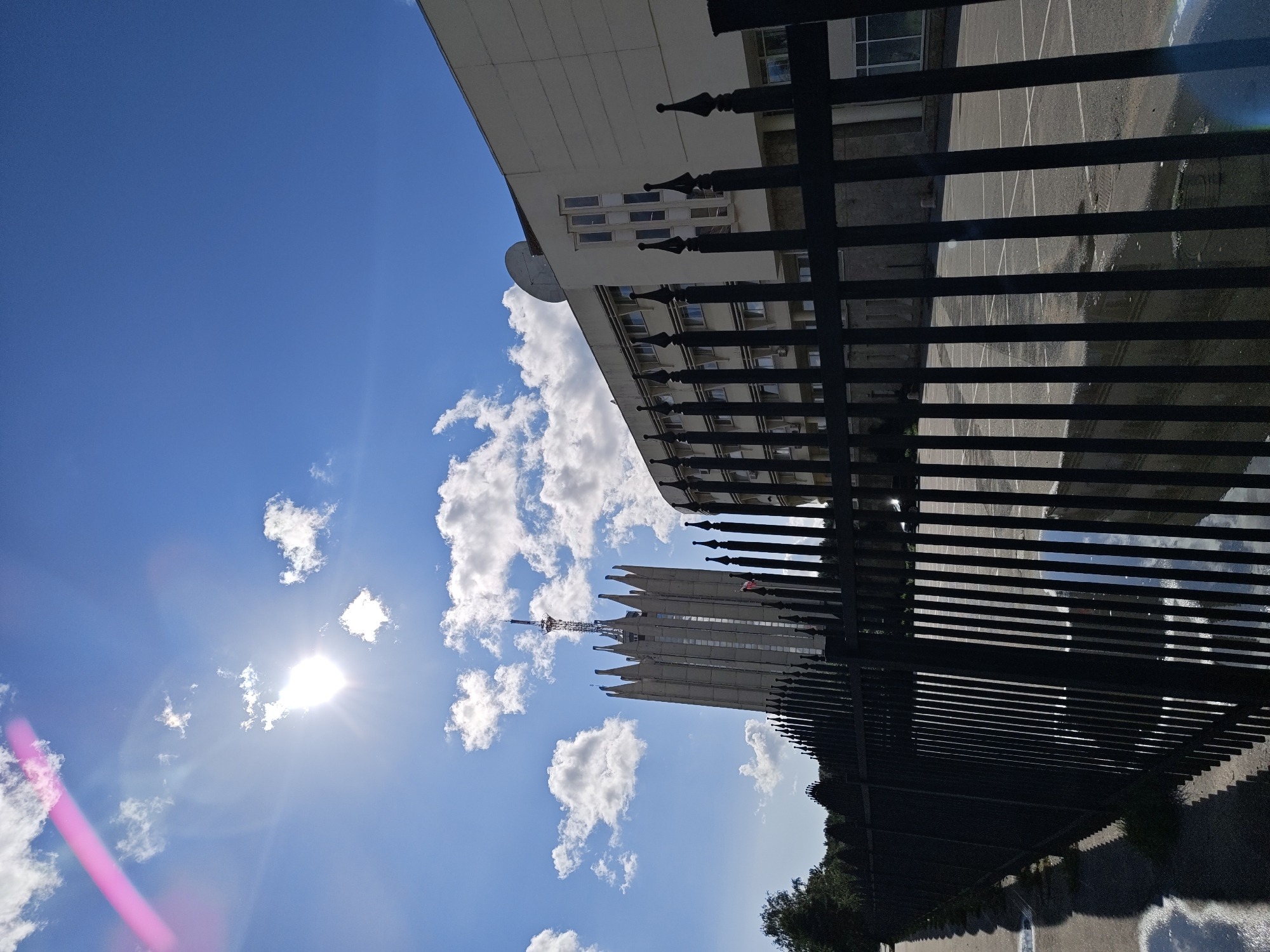 The image captures a sunny day in Saint Petersburg, Russia, with a clear blue sky and scattered white clouds. In the foreground, a black metal fence runs horizontally across the frame, with its sharp, pointed spikes casting shadows on a wet asphalt surface. Beyond the fence, two buildings are visible. To the left, a tall, imposing structure with a pointed roof and a prominent antenna stands against the sky. To the right, a lighter-colored, modern building with multiple windows and a satellite dish on its roof is partially obscured by the fence. The sun is bright, casting lens flares and creating reflections on the wet ground. The scene appears to be an outdoor area, possibly a street or a courtyard, with some greenery visible in the background.