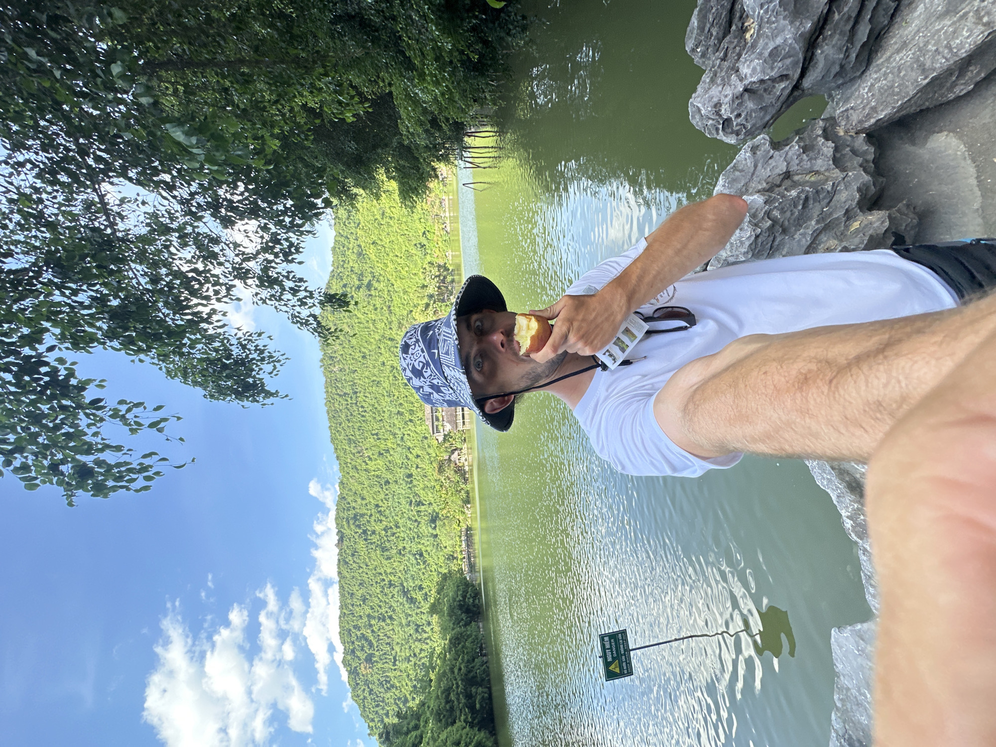 A man is taking a selfie while standing by a lake in Yên Sơn Ward, Vietnam. He is wearing a white t-shirt, black shorts, and a patterned bucket hat. He is biting into a piece of fruit. The lake is surrounded by lush green mountains and trees. In the background, there is a sign in Vietnamese that reads "NGUY HIỂM" and "DANGER". The water is calm and reflects the sky. The man's arm is extended to take the photo, and his other hand is holding the fruit. The sun is shining, and there are some clouds in the sky.
