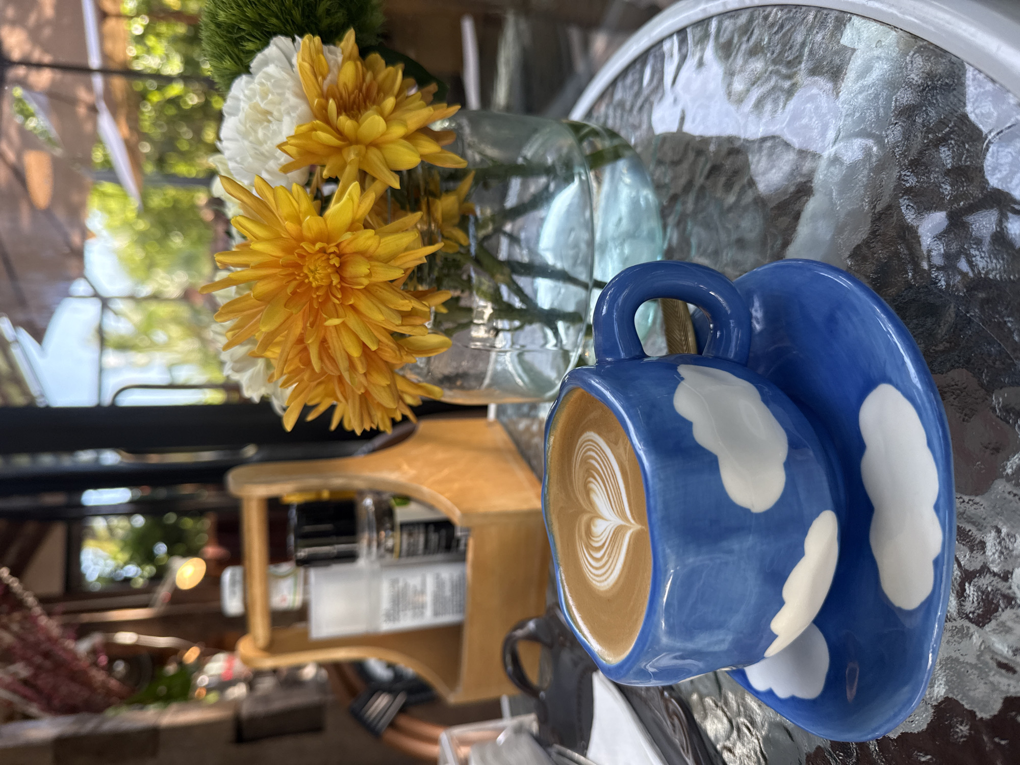 An outdoor cafe table in Ko Samui District, Thailand, displays a blue ceramic coffee cup and saucer decorated with white clouds, filled with latte art, alongside a glass vase containing yellow and white flowers. The table, with a textured glass surface, reflects the surrounding environment, which includes outdoor umbrellas and lush greenery. In the background, a wooden caddy holds various items, possibly condiments, suggesting a casual dining setting. The scene is captured in bright daylight, with sunlight creating highlights and shadows throughout the frame.