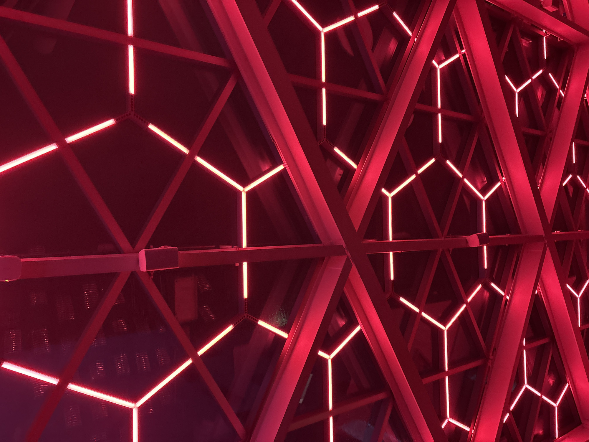 The image shows a section of the Soumaya Museum in Mexico City, Mexico, as seen from an upward angle. The architectural design features a grid of red-colored beams forming triangular and hexagonal patterns. Embedded within these patterns are glowing red linear lights, emphasizing the geometric shapes. The background is dark, contrasting with the brightly lit structure. There are no people visible in the scene. The focus is on the building's unique design and lighting.