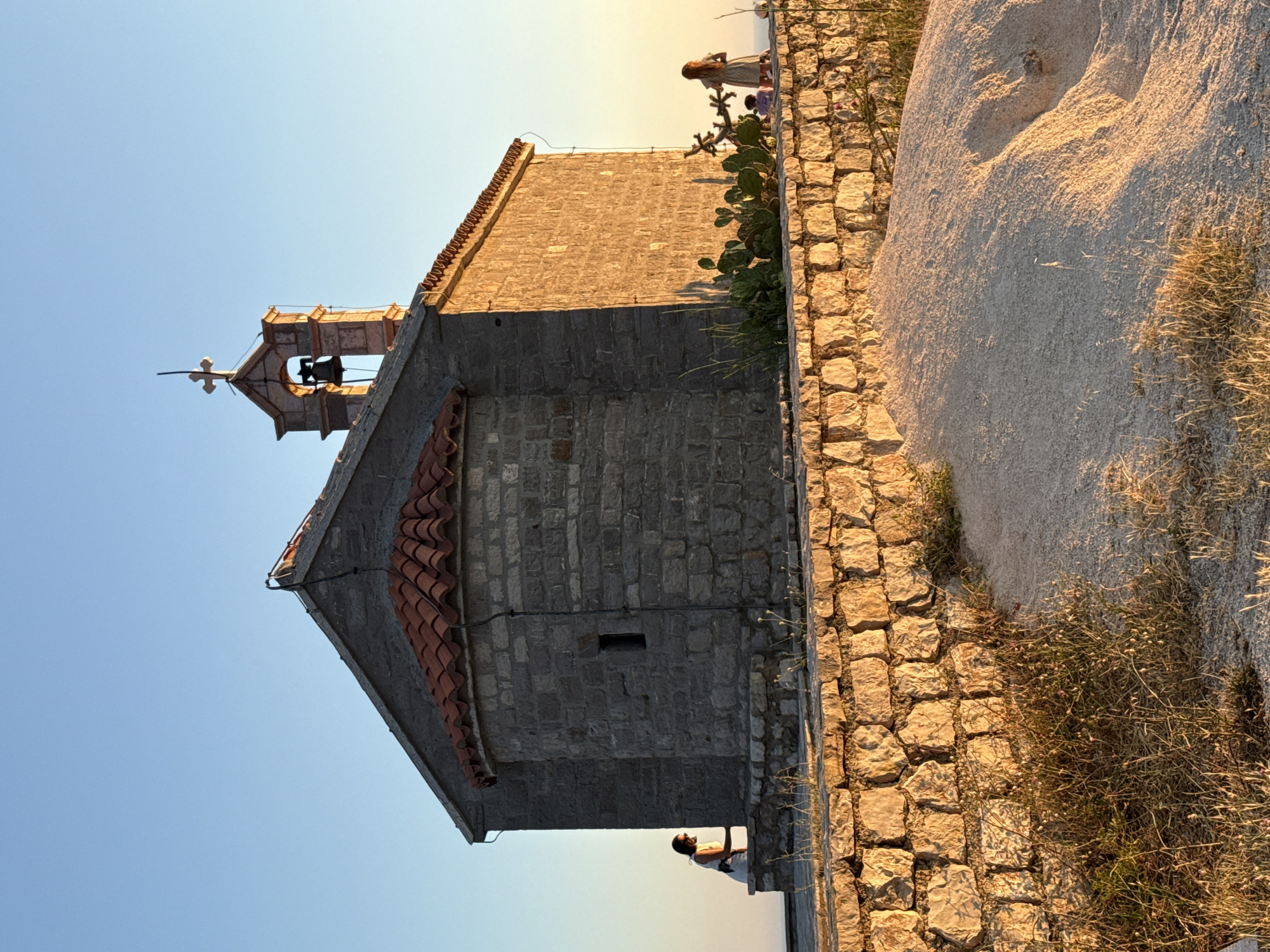 The image depicts the Church of St. John, situated on a small island off the coast of Pržno, Montenegro. The sun is setting, casting a warm, golden light on the stone structure and the surrounding landscape. The church is small and ancient, built from rough-hewn stones. Its roof is tiled with terracotta, and a simple bell tower with a cross sits atop the building. In the foreground, a low stone wall separates the church from the viewer. There are several people visible in the scene. On the left, a woman in a white top and dark pants stands leaning on the wall. On the right, another woman, possibly a child, stands near some cactus plants. In the distance, a small group of people is visible. The ground is covered with sand and sparse vegetation. The sky is clear and blue.