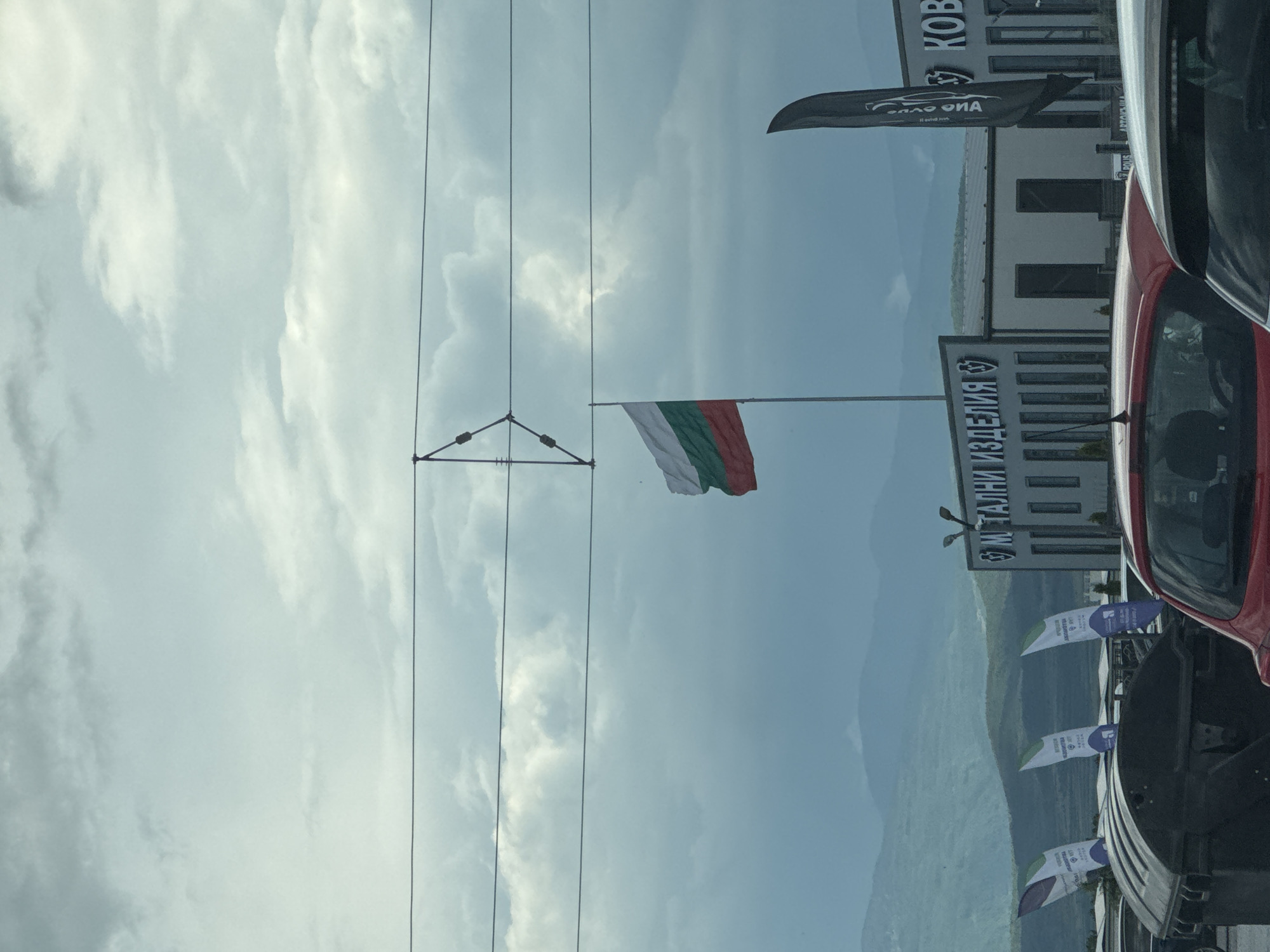 The image shows the Bulgarian flag flying against a cloudy sky. Electrical wires are visible in the foreground. In the background, there is a building with signage written in Cyrillic, partially obscured by a black flag with white text. A red car is partially visible on the right side of the image. There are multiple white and blue flags along the side of the road. Based on the Cyrillic writing and Bulgarian flag, the location is likely in Bulgaria. Based on the building signs "Метални изделия" (Metal products), and "KOBA" the location is likely the KOBA metal products shop in Plovdiv, Bulgaria.