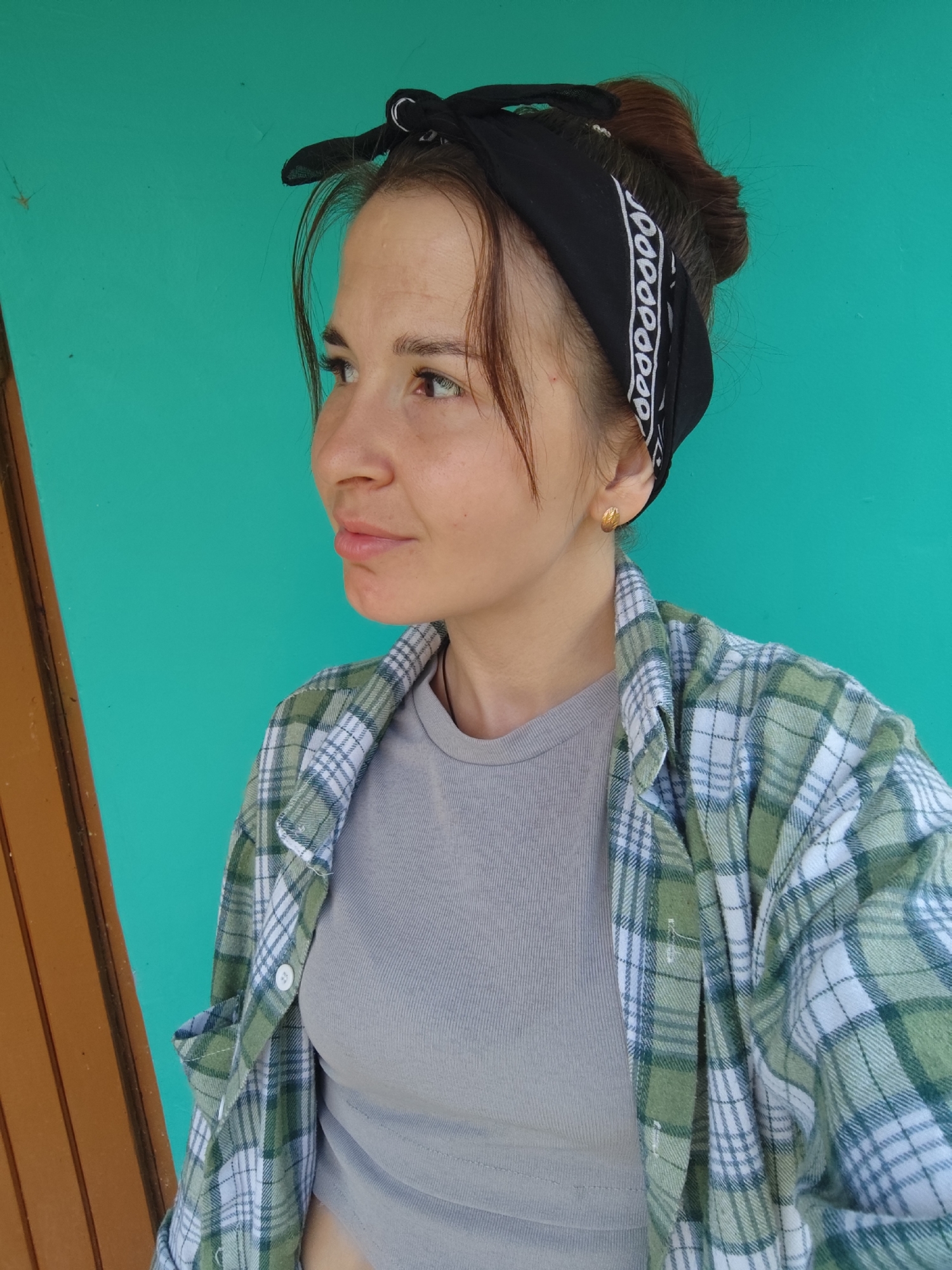 A young woman is pictured from the chest up, against a teal-colored wall. She has brown hair pulled back into a bun and is wearing a black bandana. Her head is turned to her left, and she is wearing a grey t-shirt and an unbuttoned green and white plaid shirt over it. A gold earring is visible in her left ear. To the left of the woman, a portion of a brown wooden structure is visible. The overall lighting suggests an outdoor daytime setting.