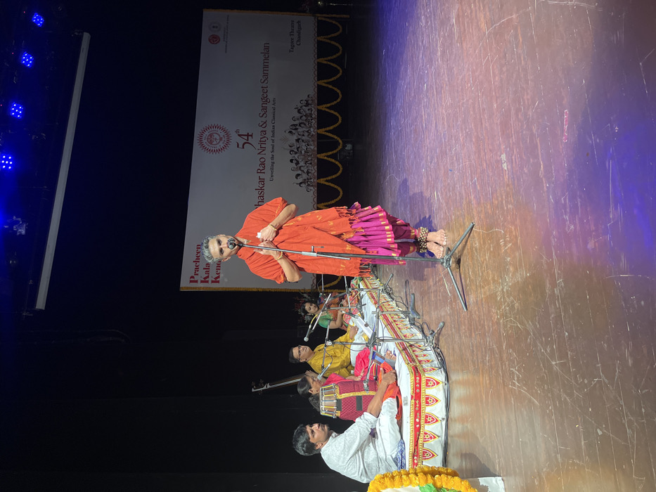 The image shows a performance at Tagore Theatre in Chandigarh, India (latitude: 30.733, longitude: 76.789). A male performer is standing center stage, wearing an orange shawl over a pink dress. He is holding a microphone and speaking. There are four musicians seated on the stage, all wearing traditional clothing. They are playing instruments, including a tabla, a sarod, and a harmonium. There are microphones in front of them, and they are holding music sheets. The background features a large banner with the words "Pracheen Kala Kendra" and "54th Bhaskar Rao Nritya & Sangeet Sammelan" on it. The stage is wooden and brightly lit. There are garlands hanging from the ceiling, creating a festive atmosphere.
