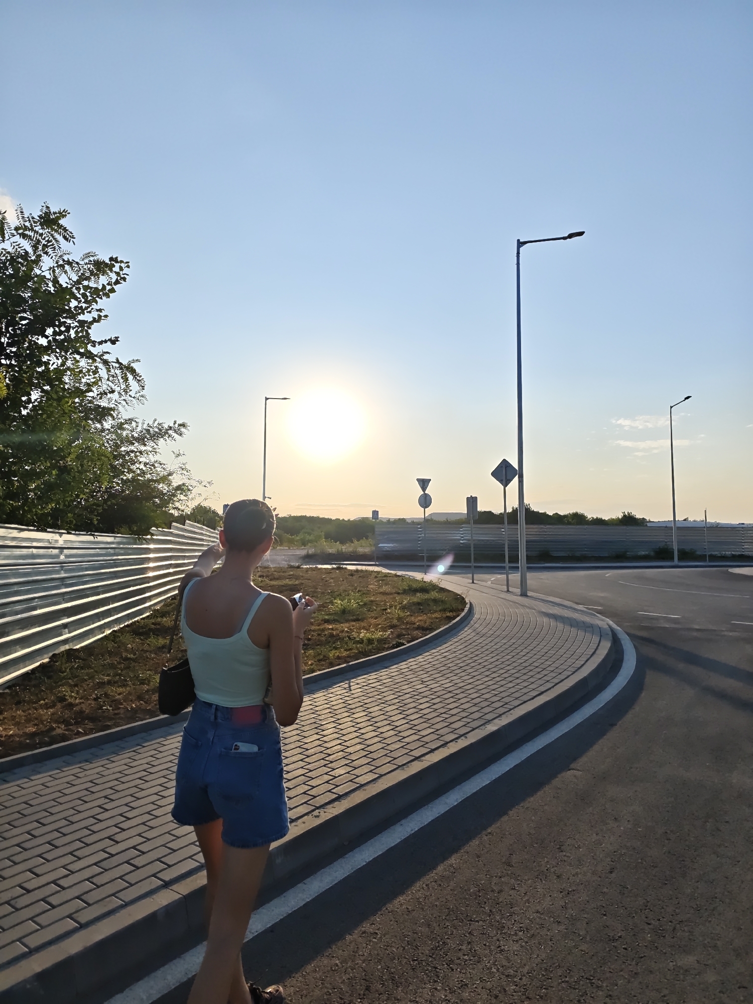 In the city of Lovech, Bulgaria, a young woman with a bun hairstyle and wearing a light blue tank top and denim shorts walks along a paved sidewalk adjacent to a road during golden hour. She is holding a mobile phone in her right hand and appears to be looking at it. The sidewalk is constructed of light brown interlocking pavers and is bordered by a grey curb. The road, paved with asphalt, curves to the right and has white lane markings. In the background, a tall metal fence stretches horizontally across the scene, with green trees and vegetation visible beyond it. Several streetlights are positioned along the road and sidewalk, and traffic signs are visible in the distance. The sun is low in the sky, casting a warm glow and long shadows, indicating either sunrise or sunset. The sky is a clear blue with a few wispy clouds.