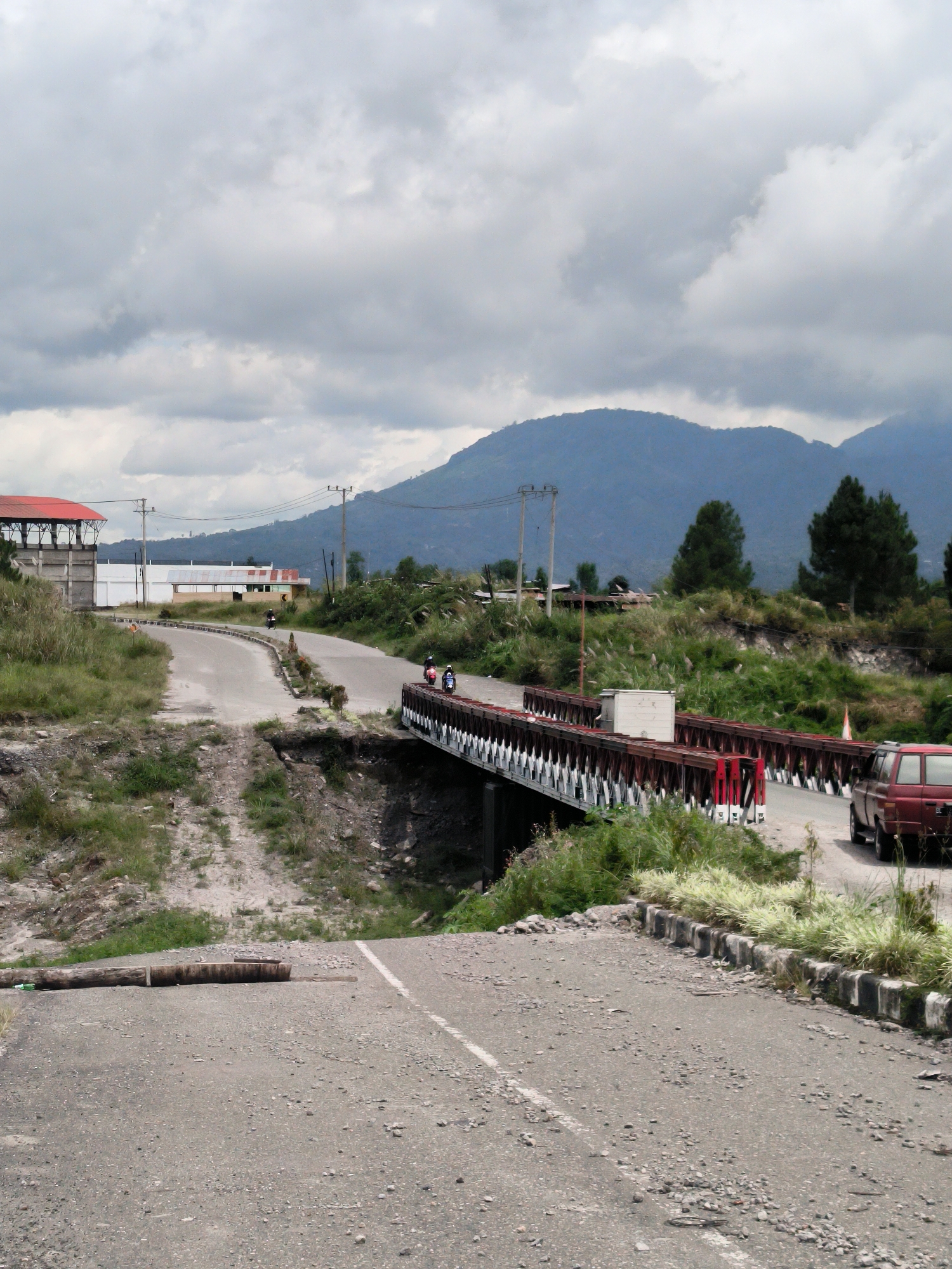 This is a vertical, medium-shot, outdoor photograph of a damaged road and bridge infrastructure in Karangrejo, Indonesia. In the foreground, a severely damaged asphalt road segment is blocked by a large, horizontally placed log. The road surface is cracked and littered with gravel and debris. To the right, a red vehicle is parked on the intact portion of the road beyond the bridge. The bridge itself is a steel truss structure with a red and white railing, spanning a dry gully. On the bridge, two individuals on a motorcycle are visible in transit, and two individuals are standing. The background features a winding asphalt road leading towards a collection of buildings, including a structure with a red roof. Beyond the buildings, lush green vegetation and trees cover the sloping terrain, which culminates in several large, blue-hued mountains under a cloudy, overcast sky. The overall scene suggests a rural or semi-rural environment impacted by damage to its transportation network.