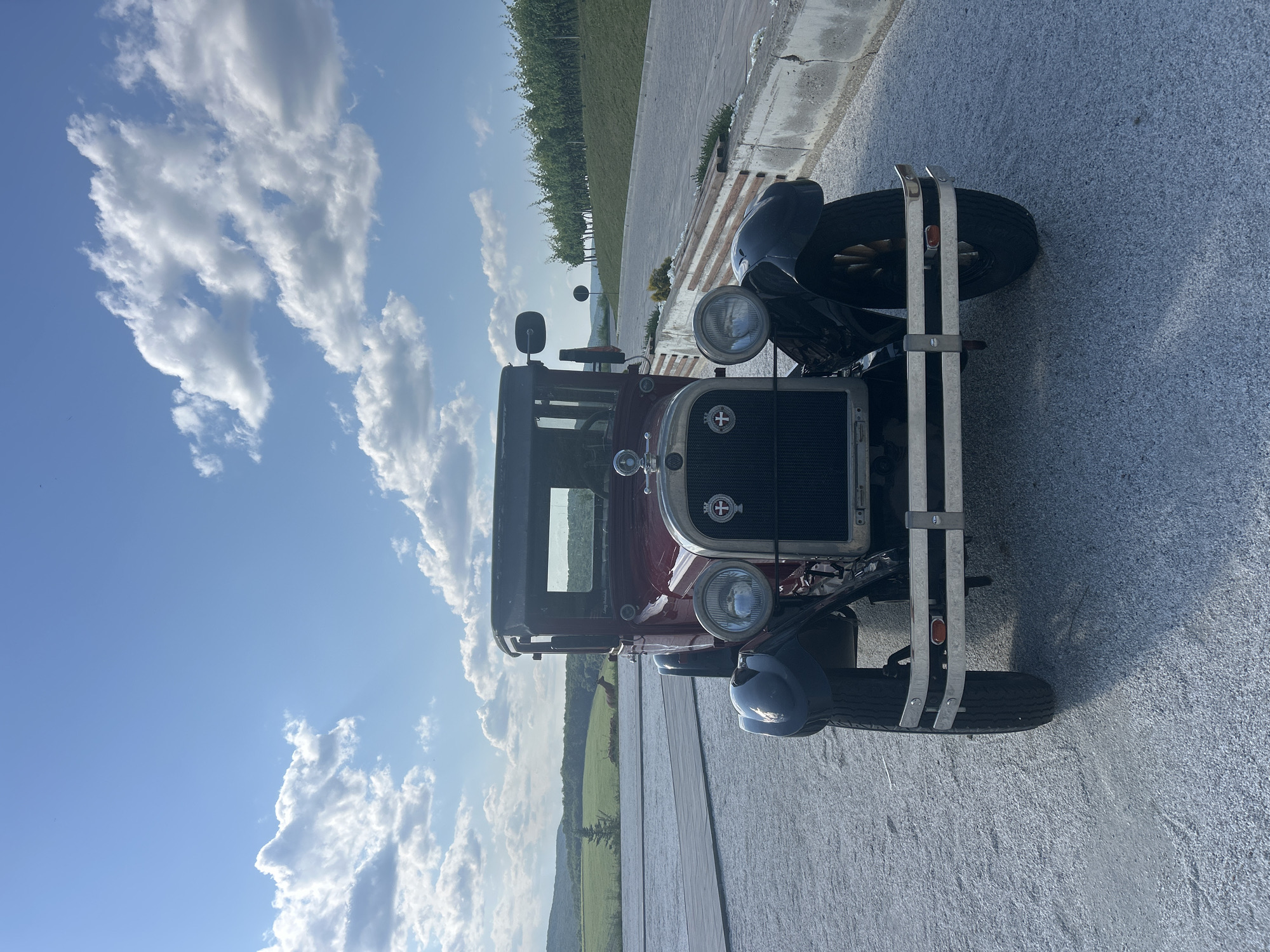 An antique red car is parked on a gravel surface in Ulmu, Moldova. The car is facing forward and is the central subject of the image. The background features a paved pathway, a low stone wall with wooden accents, a vineyard on a hillside, and rolling green hills under a blue sky with scattered white clouds. The lighting suggests it is daytime, likely late afternoon or early evening. There are no people visible in the image.