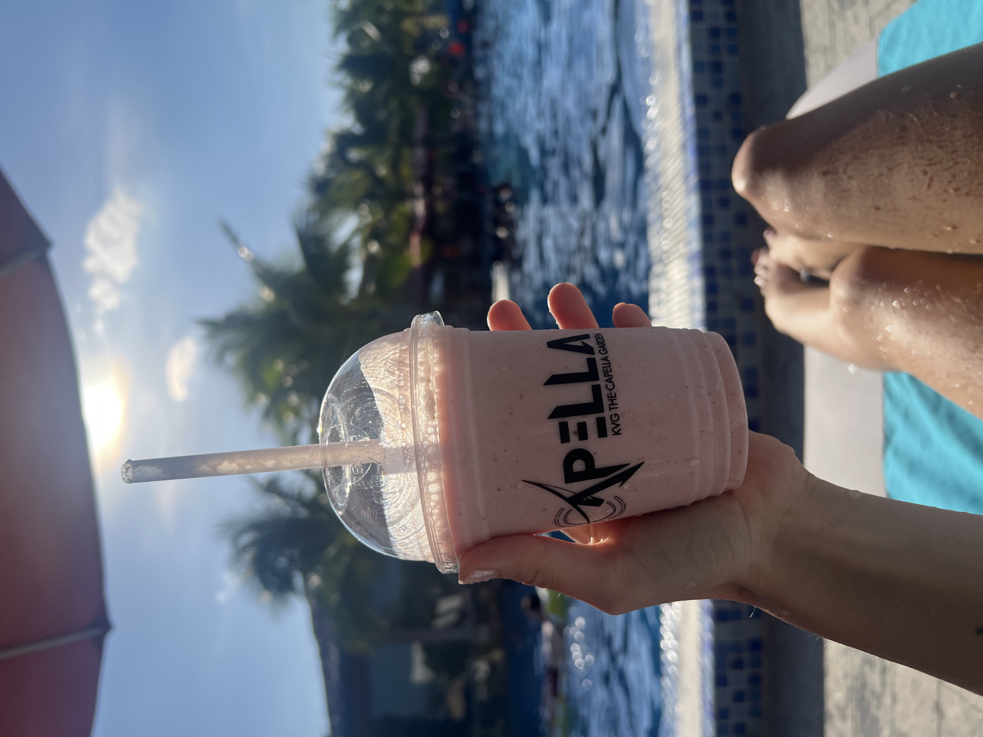 A sun-drenched outdoor scene depicts a person's perspective poolside at what appears to be a resort. The location is indicated on the cup in the person's hand as KVG The Capella Garden, which identifies the scene's location as Nam Nha Trang Ward, Vietnam. The person is sitting by a pool with legs outstretched on a light blue towel laid on a white lounge chair. The pool water is clear and blue, reflecting the sunlight. The person's hand holds a pink smoothie in a clear plastic cup, with a clear straw. The cup has a black logo on it that reads "APELLA KVG THE CAPELLA GARDEN" vertically on the side. The sun is very bright, positioned in the top left corner of the frame, and some palm trees are visible across the pool, as well as some other people partially obscured in the background.