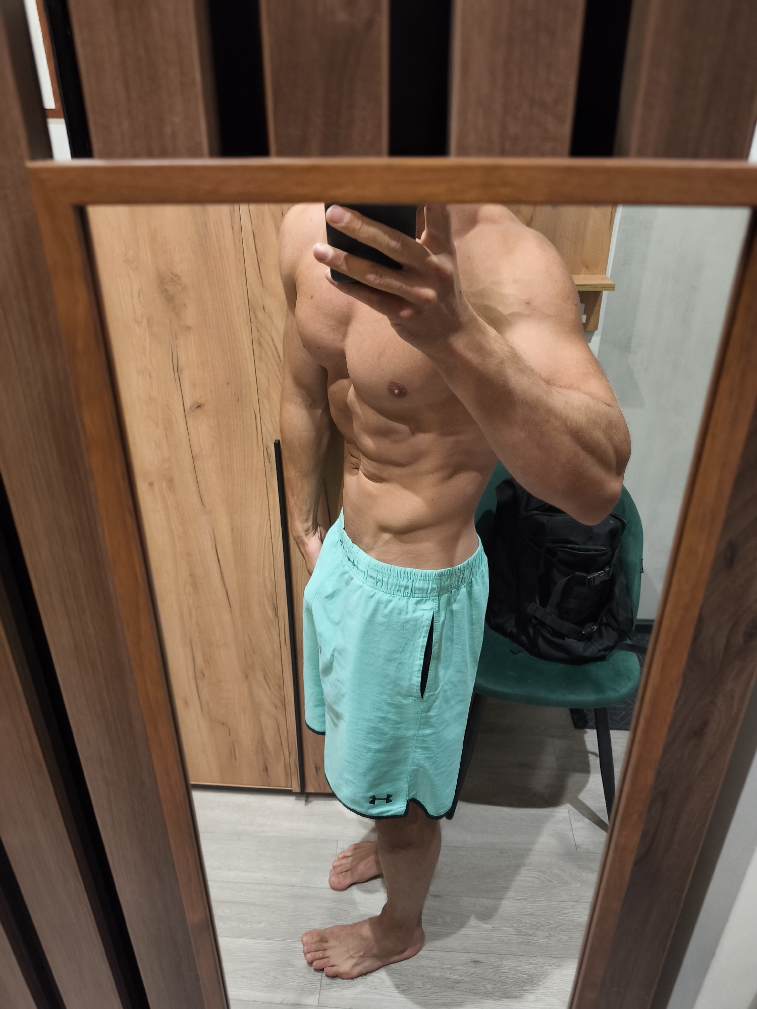 A shirtless man in light blue shorts with a black Under Armour logo is taking a mirror selfie. He is holding a black smartphone. He is standing in front of a wooden wardrobe. There is a green chair and a black backpack visible in the background. The floor appears to be light gray. The location of the city of Moscow, Russia is not discernible from the image.