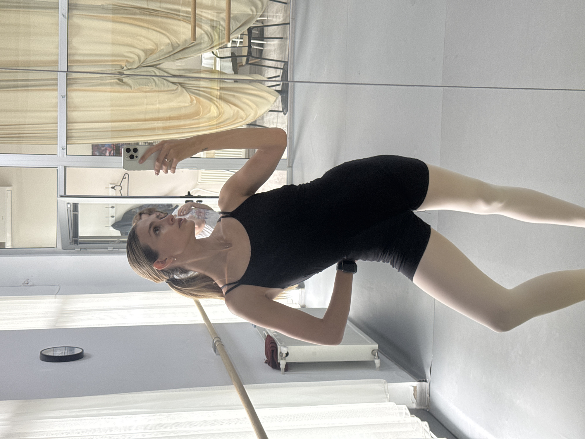 An image captures a woman, likely a ballet dancer or student, taking a mirror selfie in what appears to be a dance studio. She is positioned centrally, viewed from the waist up, and is dressed in a black form-fitting leotard or bodysuit and light-colored tights. Her hair is pulled back in a ponytail, and she wears a smartwatch on her left wrist and a ring on her right hand. She holds a light-colored iPhone, facing it towards the mirror to capture her reflection. Her gaze is directed slightly upwards, possibly at her phone screen or her reflection.
The setting is characterized by a long wooden barre that runs horizontally across the lower mid-ground, typical of a ballet studio. The wall to her left features a small, round, dark-rimmed clock. The backdrop is primarily the reflection in the large mirror, which shows windows with light-colored, possibly beige or cream-colored, curtains or blinds. Beyond these windows, vague shapes of furniture, including chairs, and an air conditioning unit are visible in the reflection, hinting at an adjacent room or outdoor space. The overall lighting is soft and natural, suggesting it is daytime. No readable text is discernible in the image. The image's context is Tbilisi, Georgia.