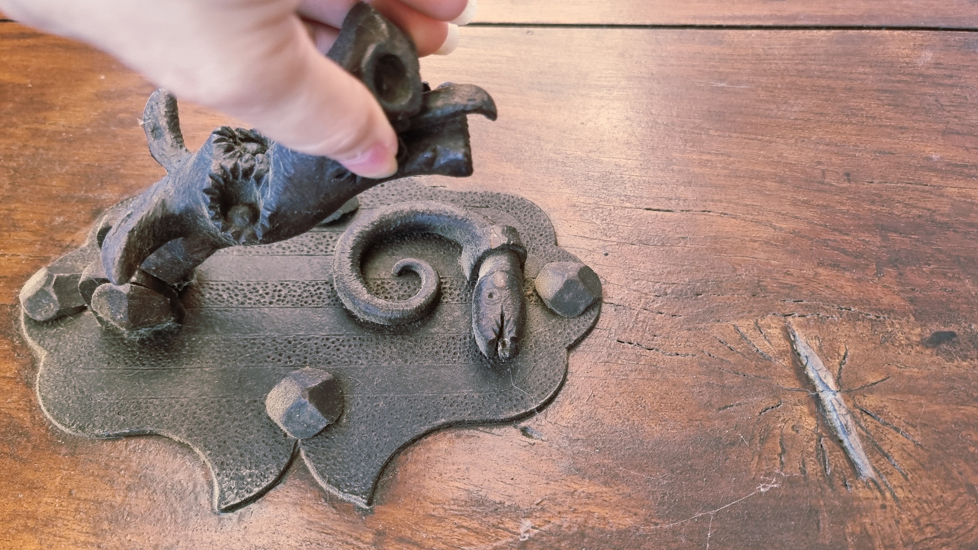 A close-up, eye-level shot captures a hand reaching for an ornate, dark metal door knocker on a rustic wooden door in Rupit, Spain. The door knocker is fashioned in the shape of a dragon or mythical creature, with its body intricately coiled and forming a snake-like head with an open mouth. The hand, belonging to a person with fair skin and short fingernails, is positioned to grasp the head of the creature. The wooden door exhibits a weathered texture with visible grain and a distinct knot with radiating cracks, suggesting age and character. The lighting is natural and casts subtle shadows, highlighting the detailed craftsmanship of the knocker and the texture of the wood.
