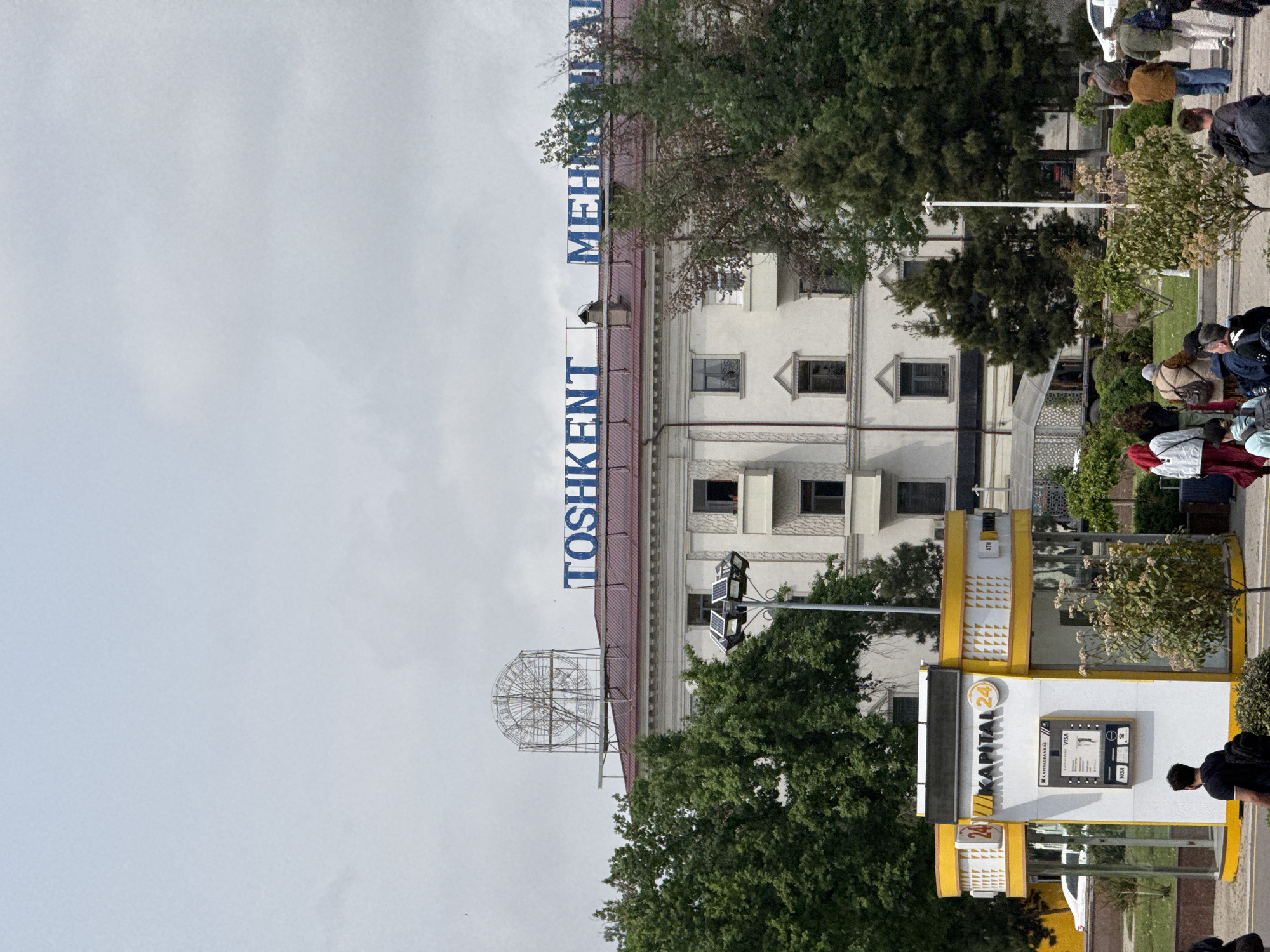 This image shows a building with the word "TOSHKENT" in large, blue vertical letters. Above that, the word "MEH" is also vertically displayed. Adjacent to the building, there is an ATM kiosk of "KAPITAL 24". The surrounding environment includes trees, other shrubbery, and people. The location is likely within the city of Tashkent, Uzbekistan.