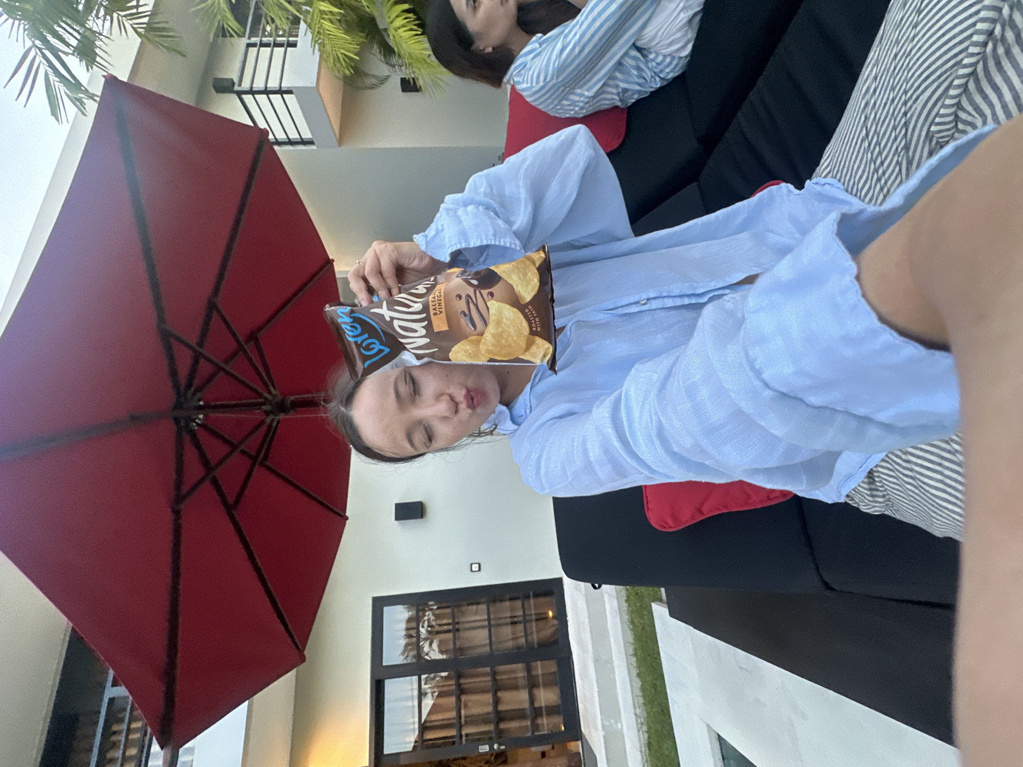 The image shows a woman holding a bag of Lays Natural Balsamic Vinegar chips, seemingly posing for a photo. She is wearing a light blue button-down shirt and appears to be reclining on outdoor furniture with black cushions and red throw pillows. Another woman is visible in the background, wearing a striped shirt and also reclining on similar furniture. A red patio umbrella is prominently featured in the foreground. The backdrop includes a building with white walls, black-framed windows, and a small palm tree. Based on the details such as the building style and vegetation, the location is likely in North Kuta, Indonesia.