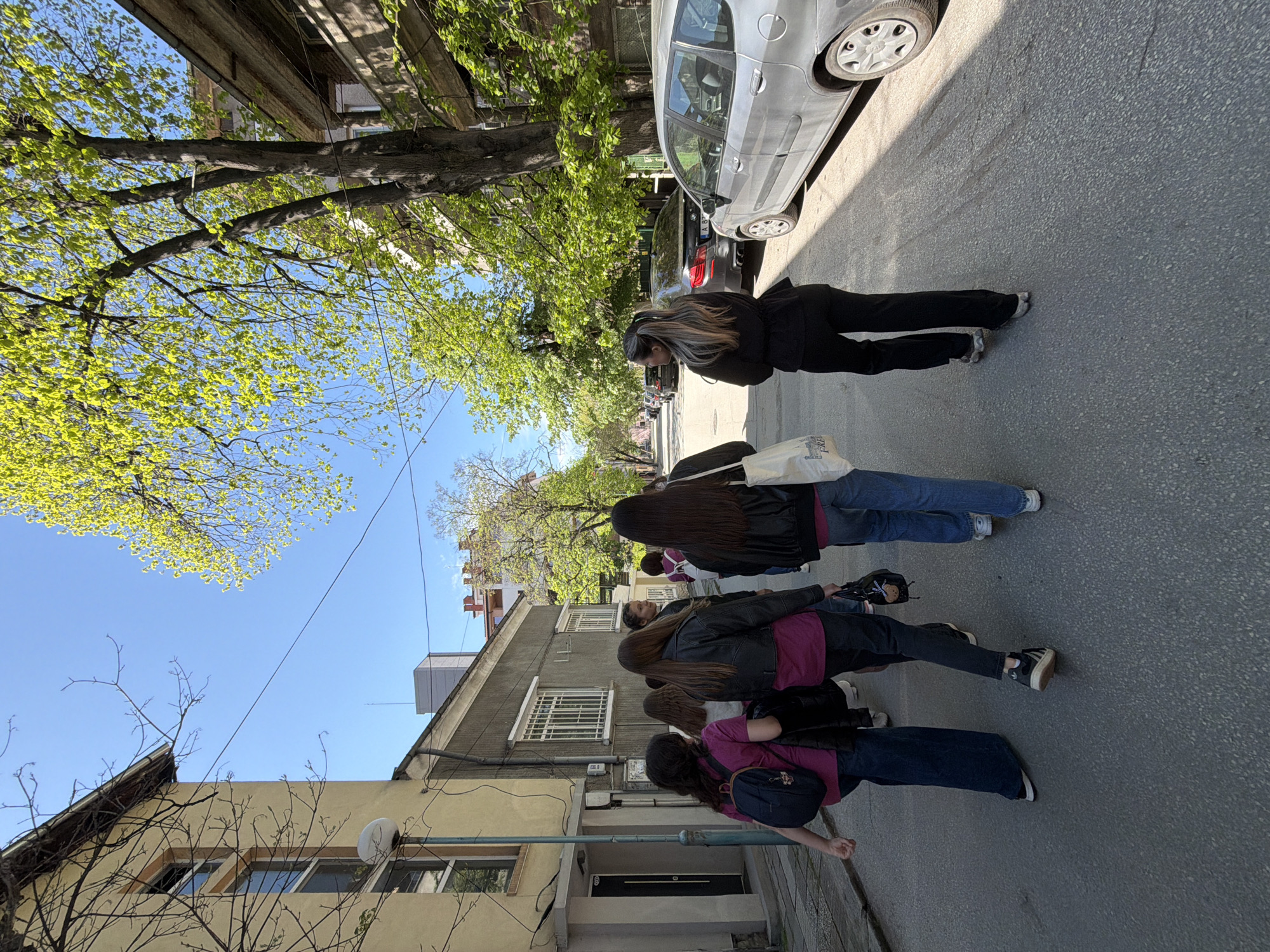 The photo depicts a group of approximately four young women walking down a street in Plovdiv, Bulgaria. They are positioned single-file on the right side of the road, near parked cars. The women are dressed casually, with jeans and jackets visible. A taller woman is walking ahead of them, taking photos. The street is lined with buildings of varying heights and architectural styles, with the blue sky and trees with green leaves overhead.
