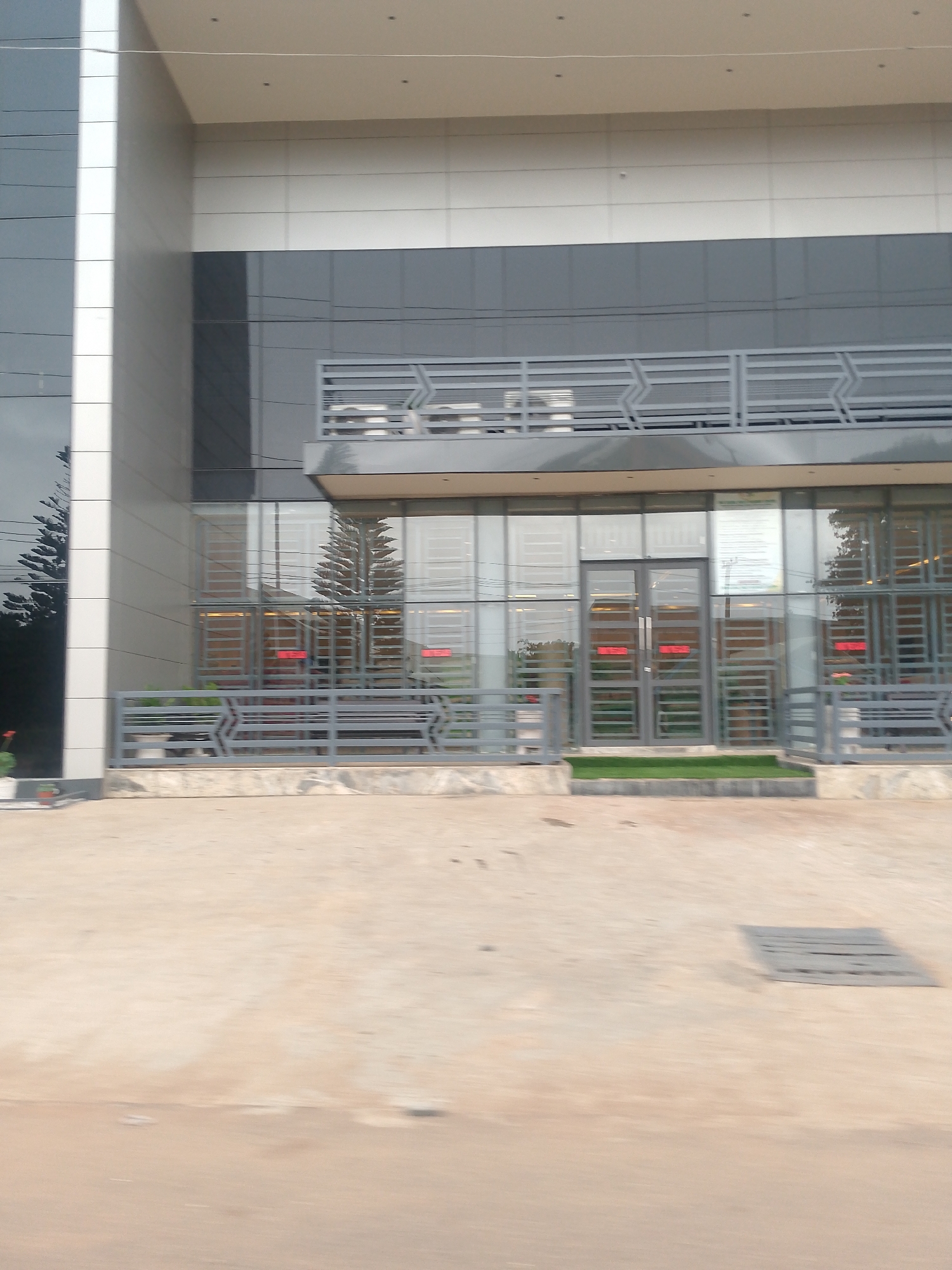 The image shows the exterior of a modern-looking building in Bukuru, Nigeria. The building has a glass facade with metal railings and a balcony. A small patch of green grass is visible near the entrance. The foreground consists of a concrete ground with a drain cover. The building appears to be a commercial or office space. No people are visible in the image.