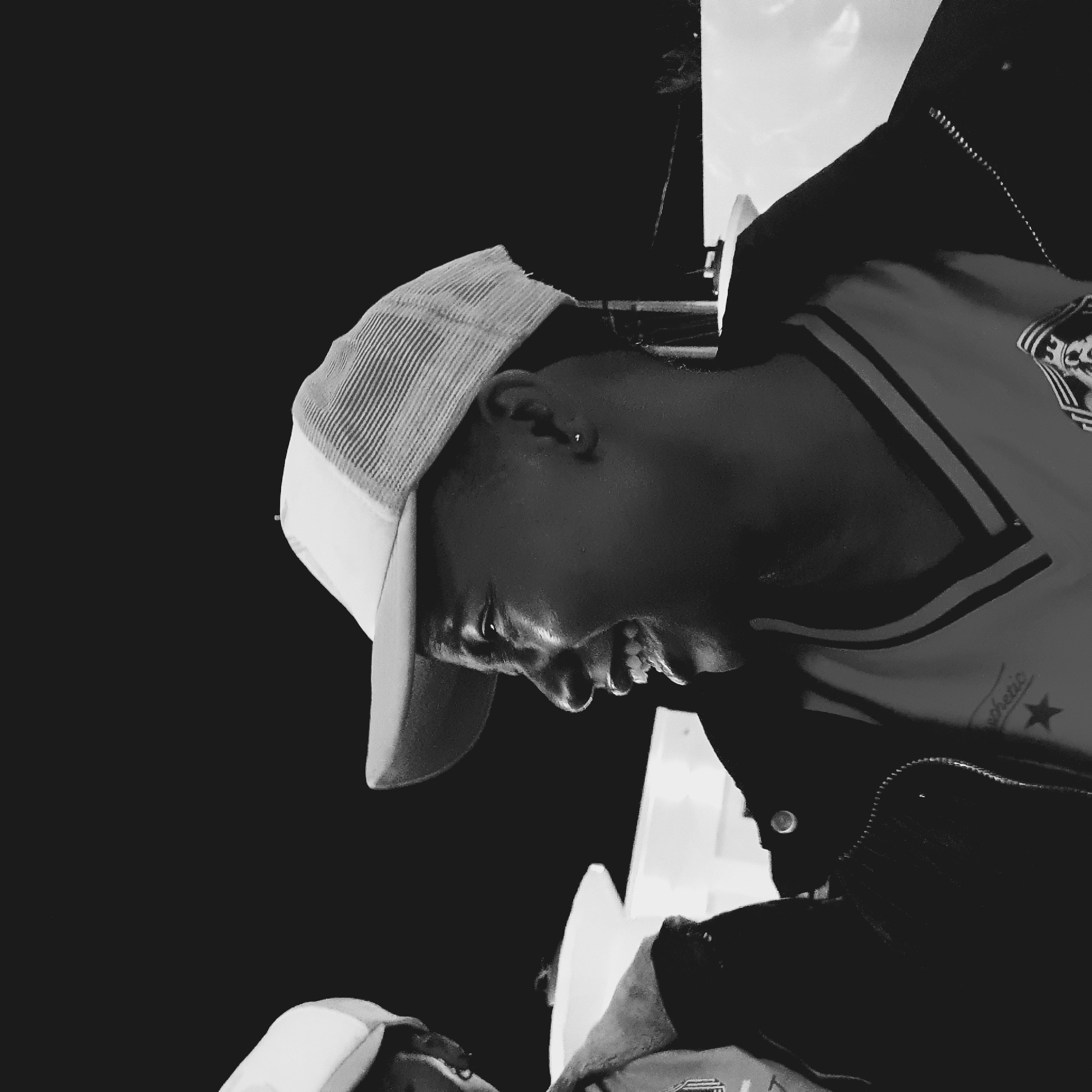 In a black and white photograph taken in Bar Jirgi Summa, Nigeria, two individuals are visible from the shoulders up, facing each other. The individual on the right, in the foreground and in focus, is wearing a mesh-backed baseball cap and a collared shirt with striped trim. They are looking to their left, with their mouth open in a wide smile, revealing their teeth. Their skin appears dark, and they are wearing a small earring in their visible ear. To the left of this person, another individual is partially visible, also wearing a baseball cap and facing towards the right. Their face is less clear, but they appear to be looking at the other person. Both individuals are wearing dark jackets over their shirts. The background is dark and appears to be outdoors at night, with some blurred white shapes suggesting structures or possibly lights in the distance. The overall lighting suggests an artificial light source illuminating the subjects.