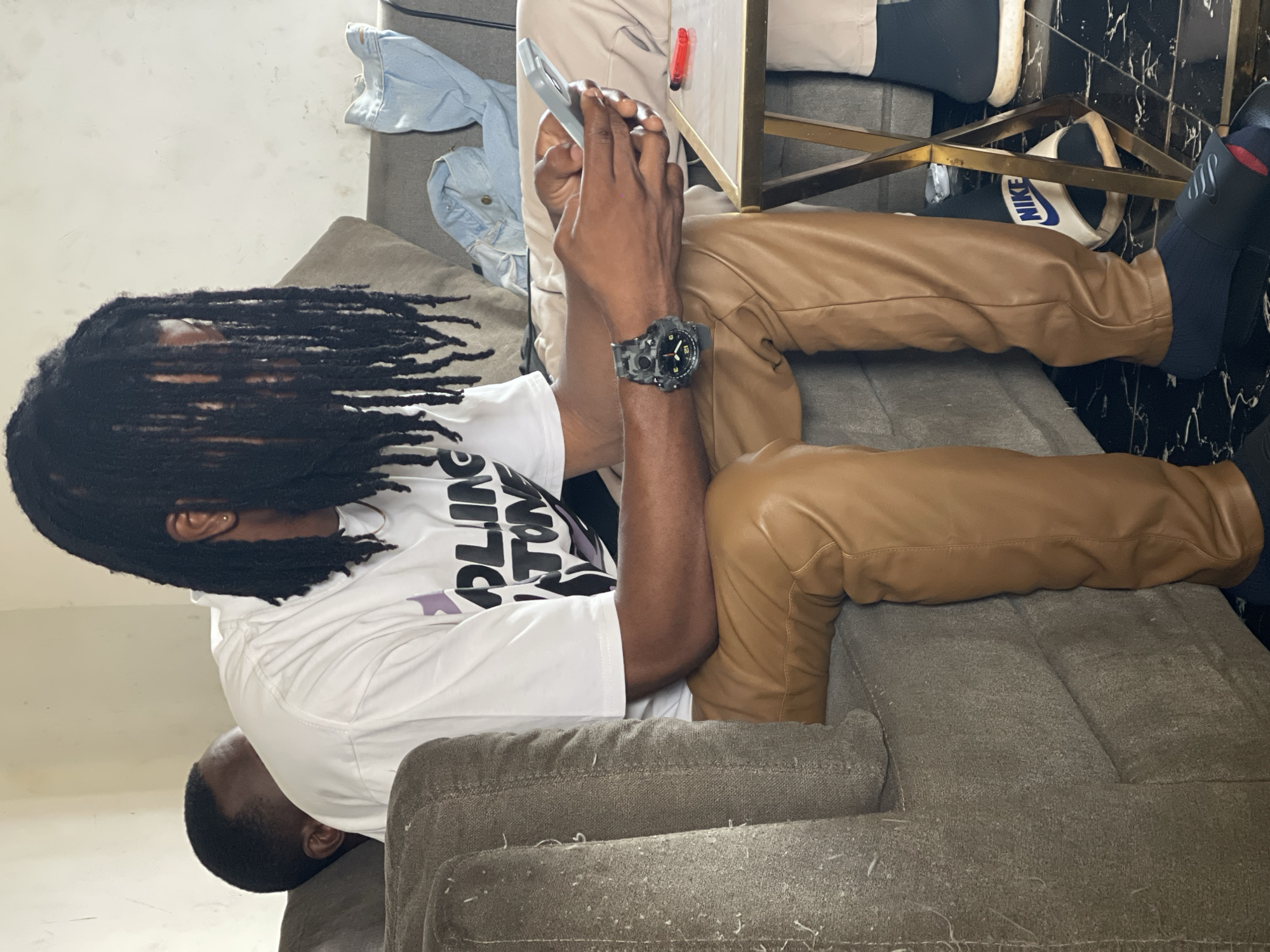 A medium shot captures two individuals in an indoor setting, possibly a living room, in Abuja, Nigeria. The primary subject, a person with dark, voluminous dreadlocks partially obscuring their face, is seated on a textured grey sofa. They are wearing a white t-shirt with dark text and brown leather-like pants. A black digital wristwatch with a camouflage pattern is visible on their left wrist. They are holding a light blue smartphone in their hands and appear to be engaged with it. To their left, another individual is partially visible, with their head turned away and resting on the sofa. In the background, folded denim clothing is present on another seating area. A small, metallic side table is situated in front of the primary subject, holding a red lighter and a partially visible phone. The floor is a black and white tiled pattern. The lighting suggests an indoor environment, possibly during the day.