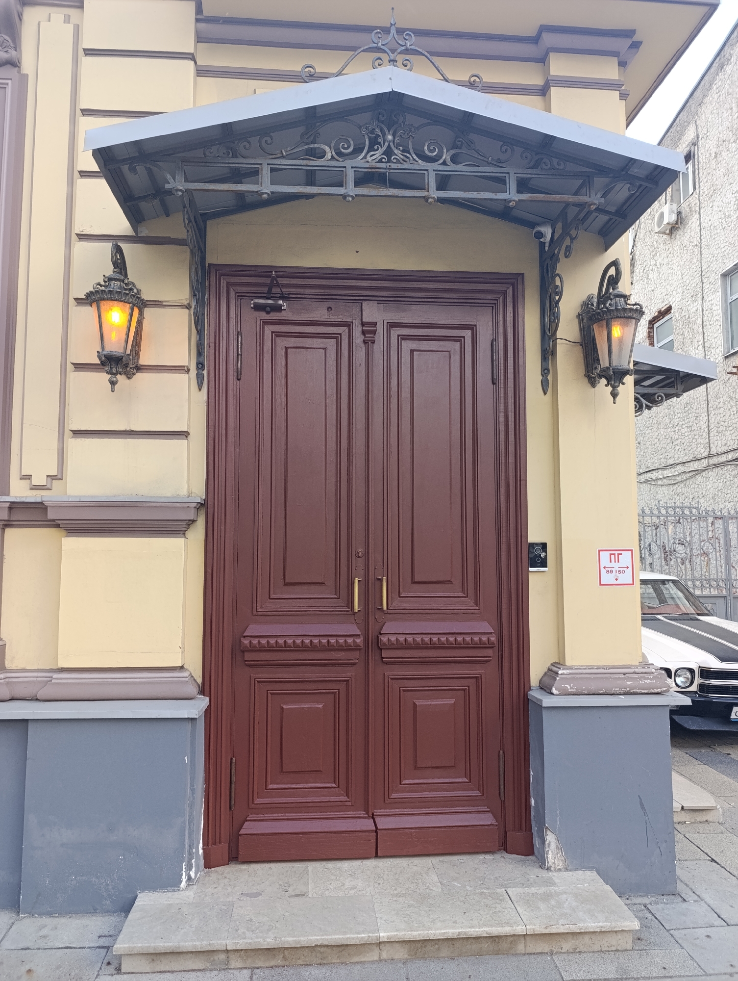A front-on, slightly low-angle shot captures the ornate entrance of a historic building, likely in Moscow, Russia. The central feature is a large, dark reddish-brown double door with intricate paneling and golden handles. Above the door, an elaborate black metal canopy with a grey roof extends, featuring decorative scrolls and supports.
Flanking the door, two ornate black metal lanterns with glowing orange lights cast a warm glow, suggesting late afternoon, dusk, or an overcast day. The building's facade is a light yellow, adorned with classical architectural details such as pilasters and cornices, set on a grey base that shows some minor wear.
To the right of the door, a small black intercom system is mounted on the wall. Below it, a red square sign with white text reads "ПГ" (likely indicating a fire hydrant location) and numbers "↔ 89 ↕ 50", which probably denote distances in centimeters. A small security camera is visible mounted under the canopy on the right side.
In the background to the right, a white car with black racing stripes is partially visible, parked on the street behind a decorative grey metal fence. Another building with windows and an air conditioning unit can be seen beyond the fence. No people are present, and the scene is static.