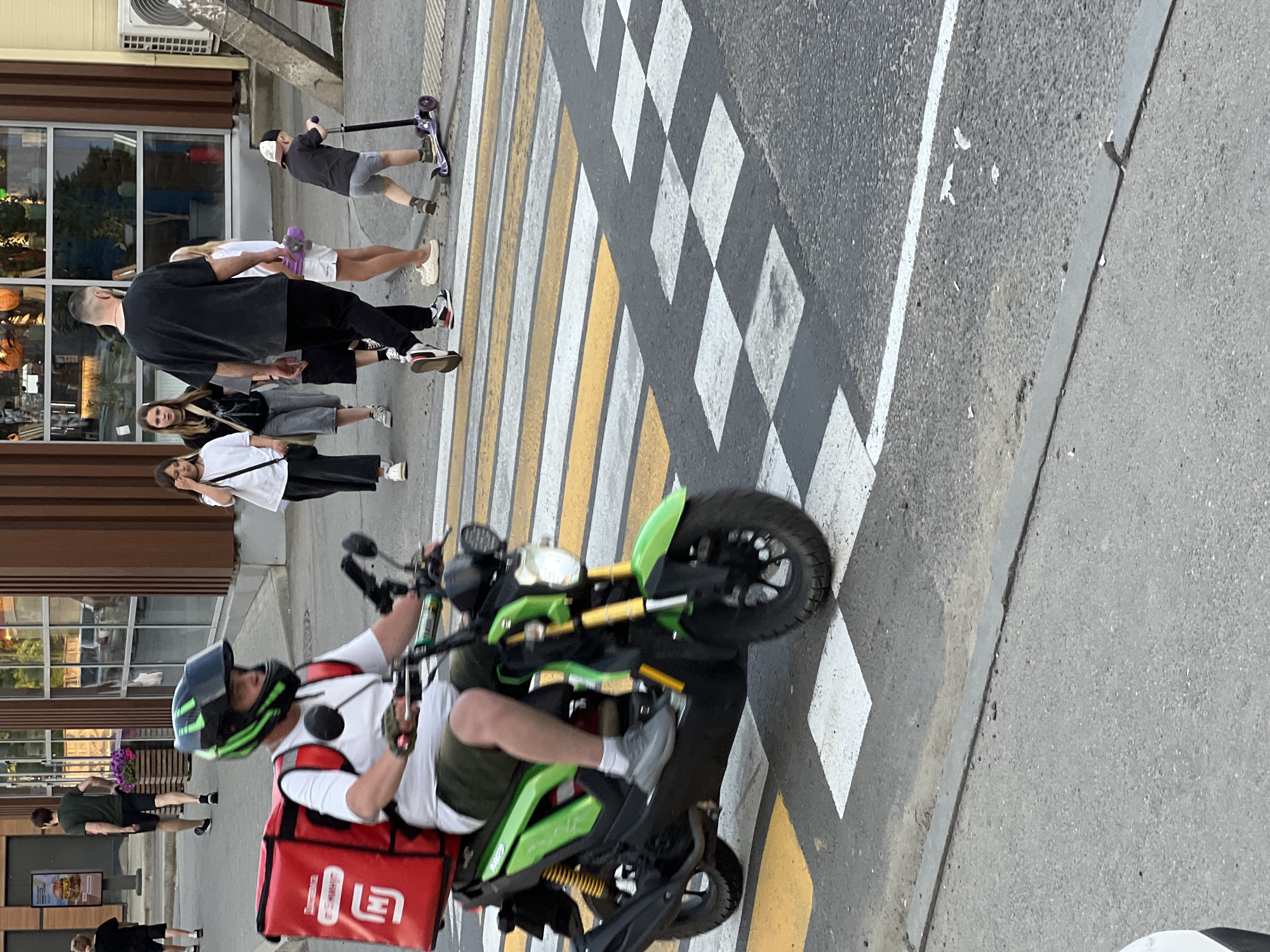 A delivery person on a green and black scooter with a large red insulated delivery bag on the back is crossing a pedestrian crosswalk. The scooter rider is wearing a white t-shirt, green shorts, grey sneakers, and a green and black helmet. In the background, several people are walking on a sidewalk next to a building with large windows. To the right, a child on a scooter is also on the crosswalk. The scene appears to be on a street in Yablonovskiy, Russia.